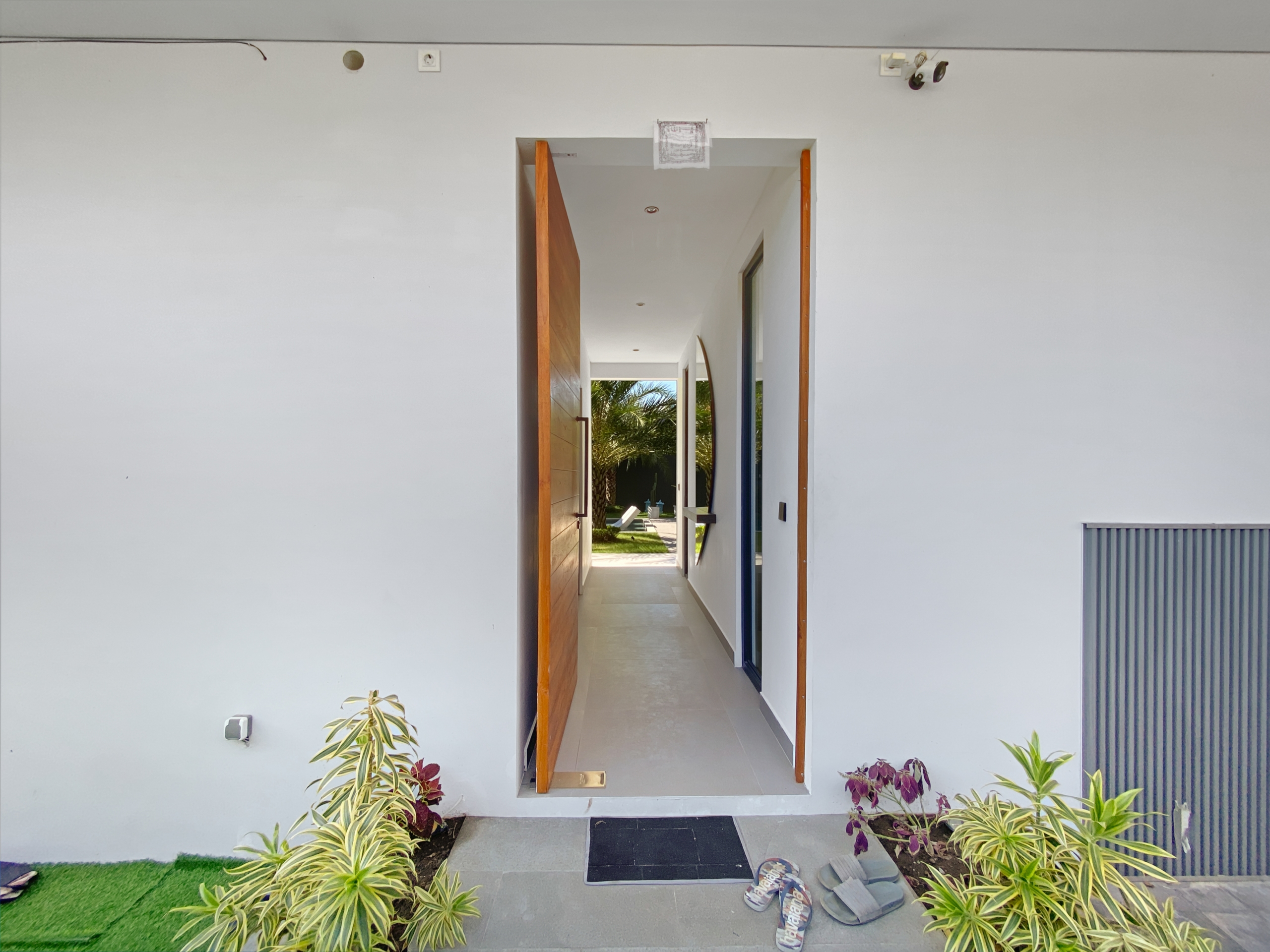 Open doorway in white wall reveals a bright hallway leading to an outdoor courtyard with palm trees; interior shows a wooden door on the left.