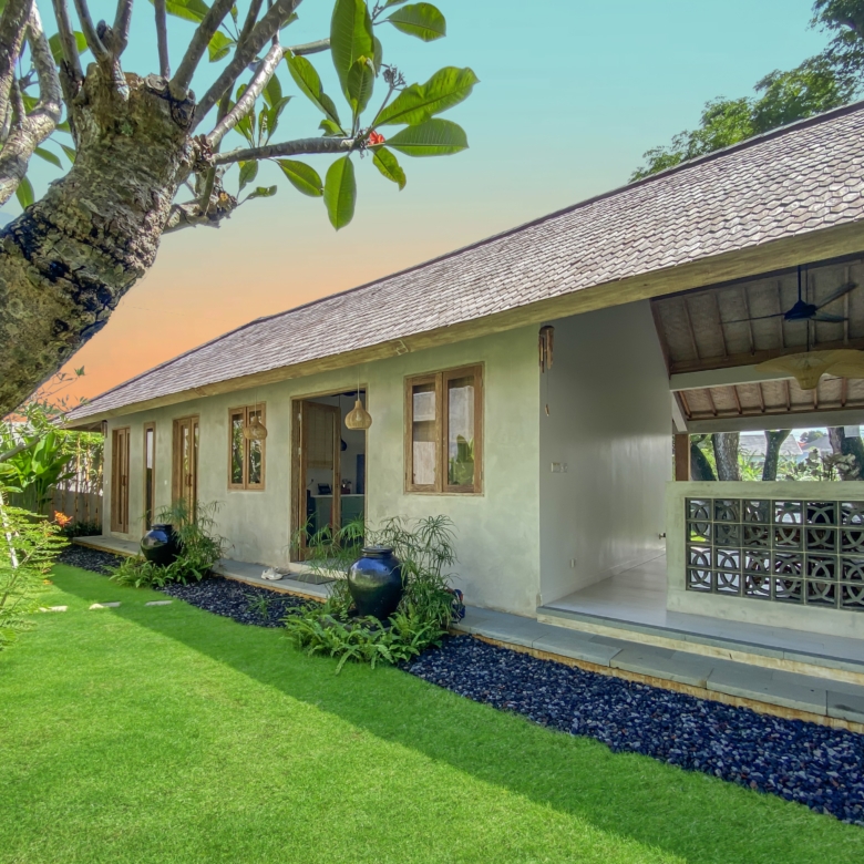 Single-story house with a sloped tiled roof, veranda, and a vibrant green lawn, with a large tree trunk in the foreground and a gravel path along the edge.