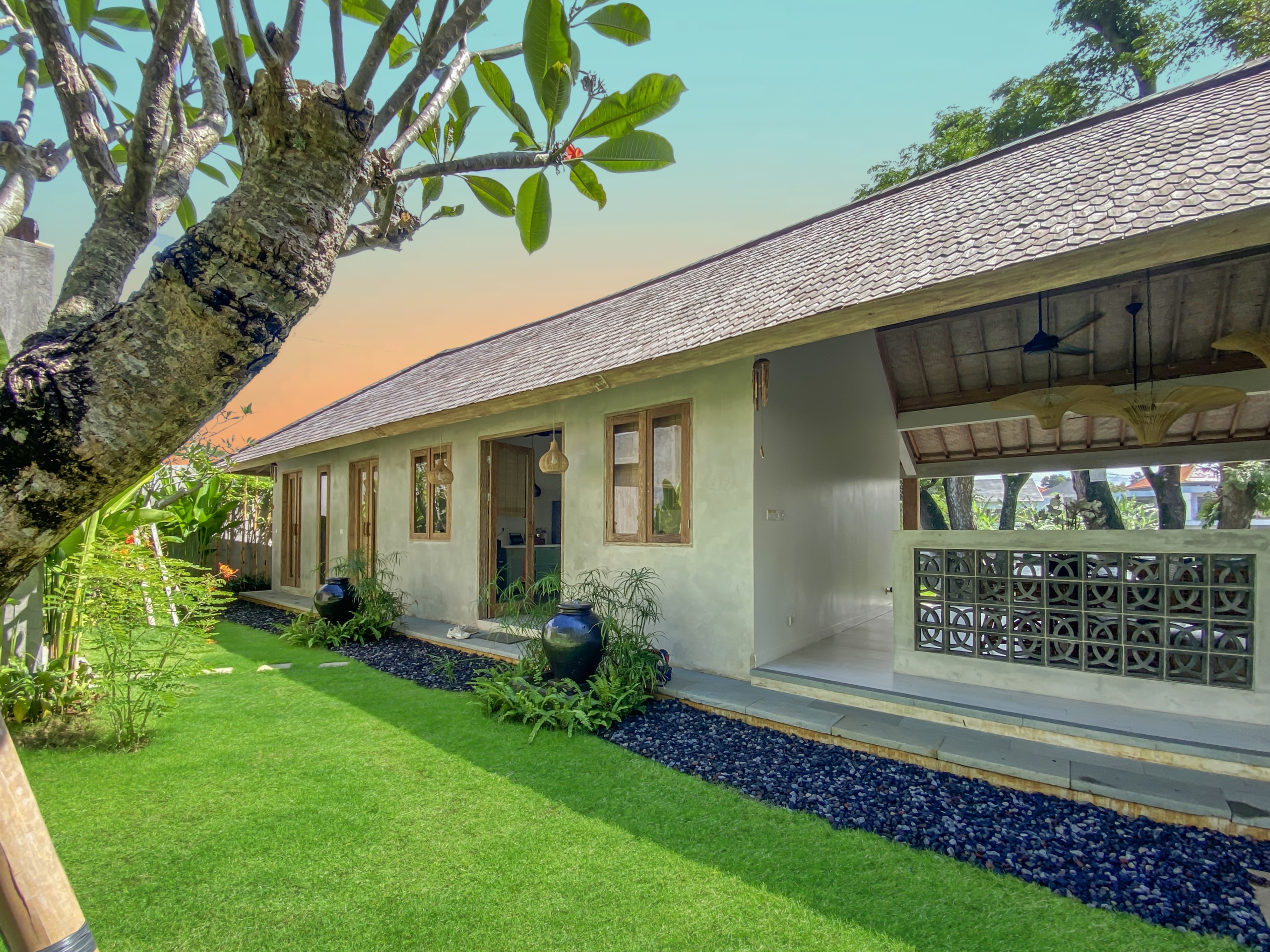 b12e6e16800d86bd8ed4e696721fff2420f2c903 Single-story house with a sloped tiled roof, veranda, and a vibrant green lawn, with a large tree trunk in the foreground and a gravel path along the edge.