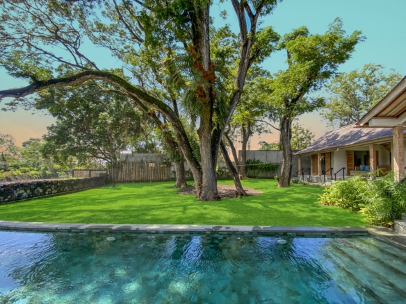 Pool in the foreground with a grassy yard and large trees casting shade, a house with a porch on the right side of the image.