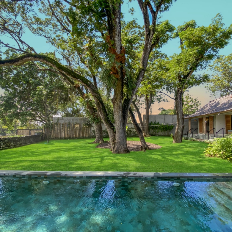 Pool in the foreground with a grassy yard and large trees casting shade, a house with a porch on the right side of the image.