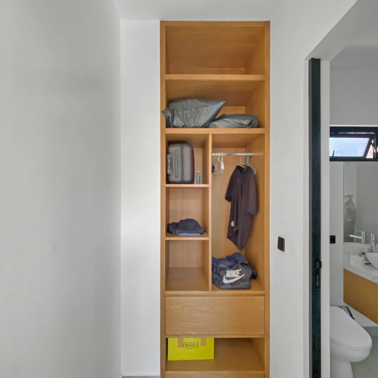 Tall wooden built-in wardrobe in a hallway with shelves holding bags, clothes, and a bright yellow 'Fragile' box on the bottom shelf; bathroom is visible to the right.
