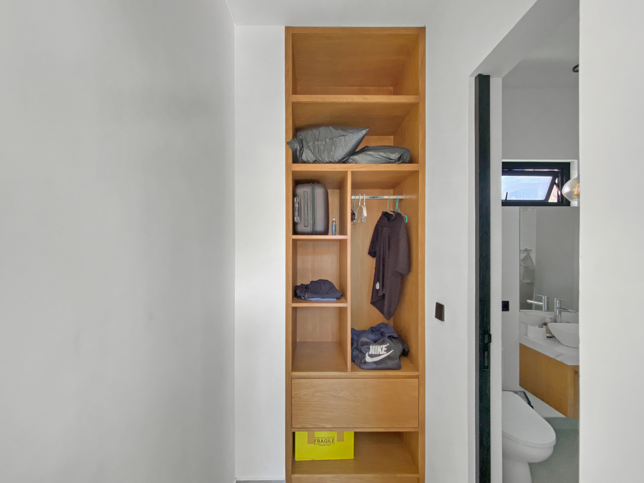 Tall wooden built-in wardrobe in a hallway with shelves holding bags, clothes, and a bright yellow 'Fragile' box on the bottom shelf; bathroom is visible to the right.