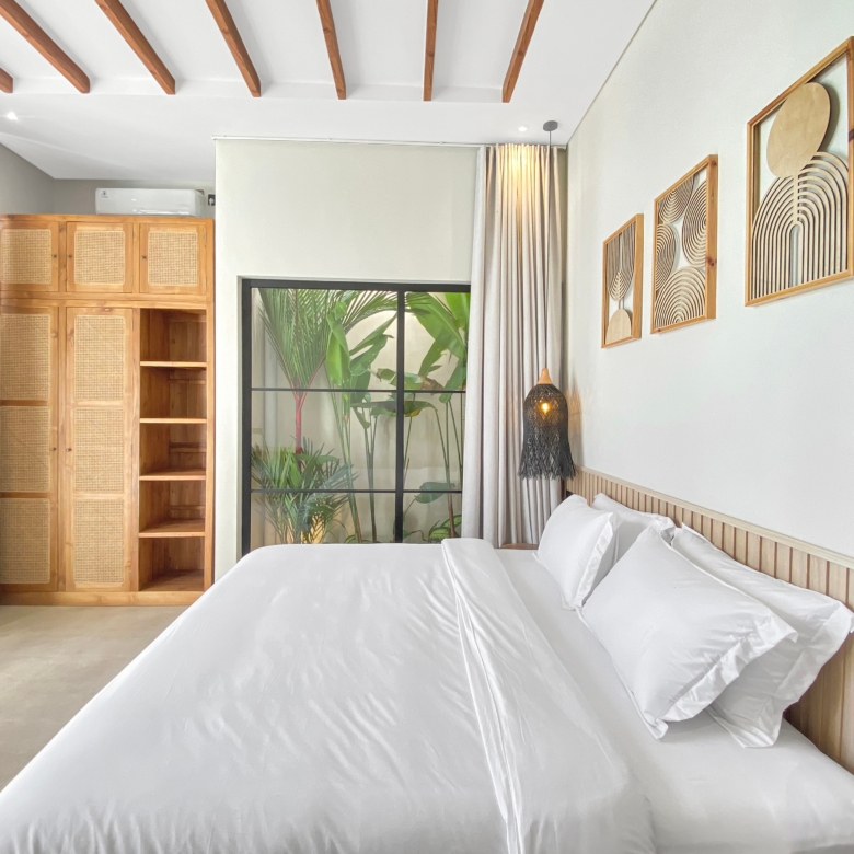 Hotel bedroom with two white-biased beds, wooden headboard, and a large window showing tropical plants outside.