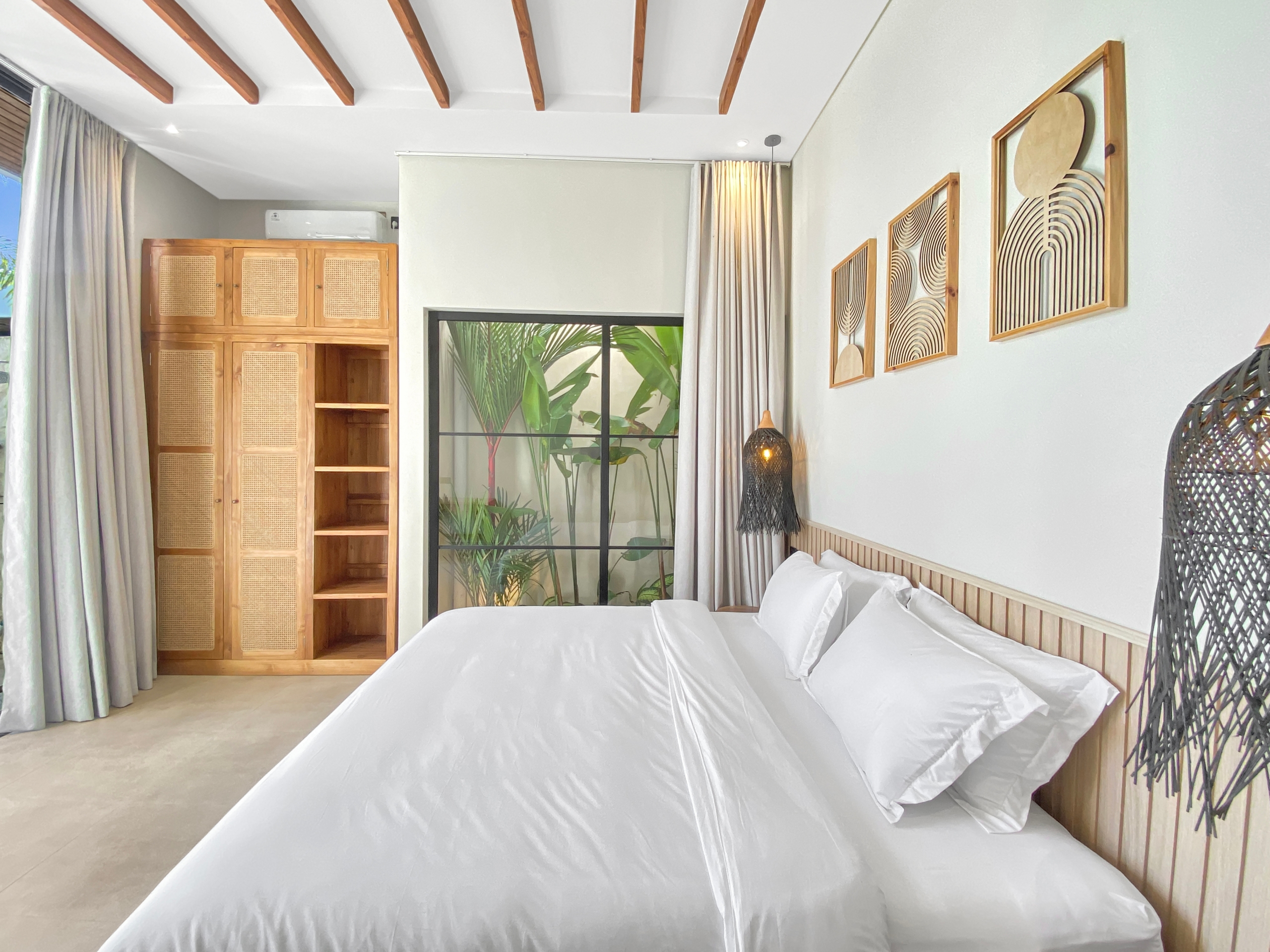 Hotel bedroom with two white-biased beds, wooden headboard, and a large window showing tropical plants outside.