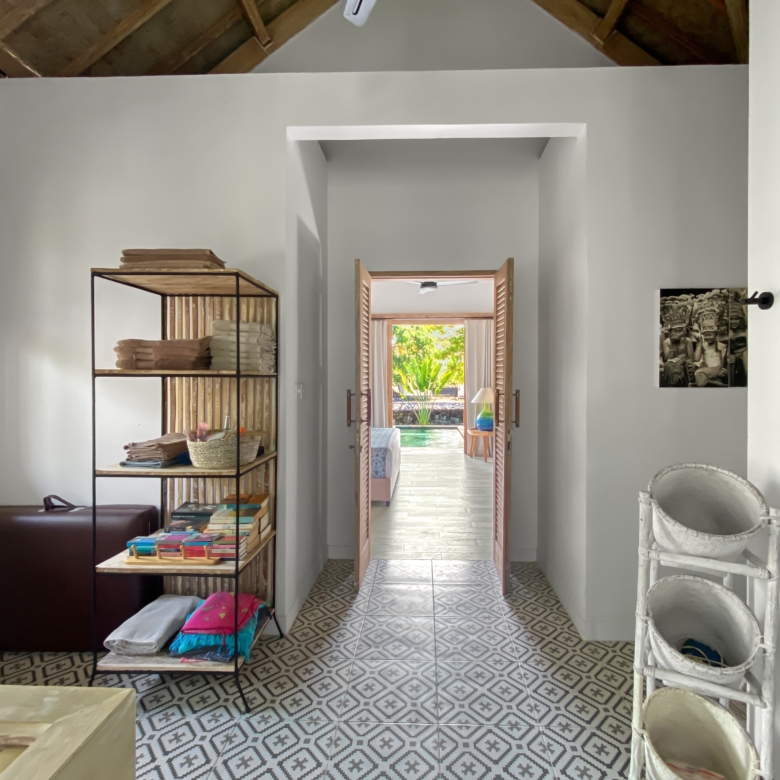 Interior view of a bright room with a metal shelving unit holding towels and fabrics, leading to a sunlit doorway to an outdoor pool area.
