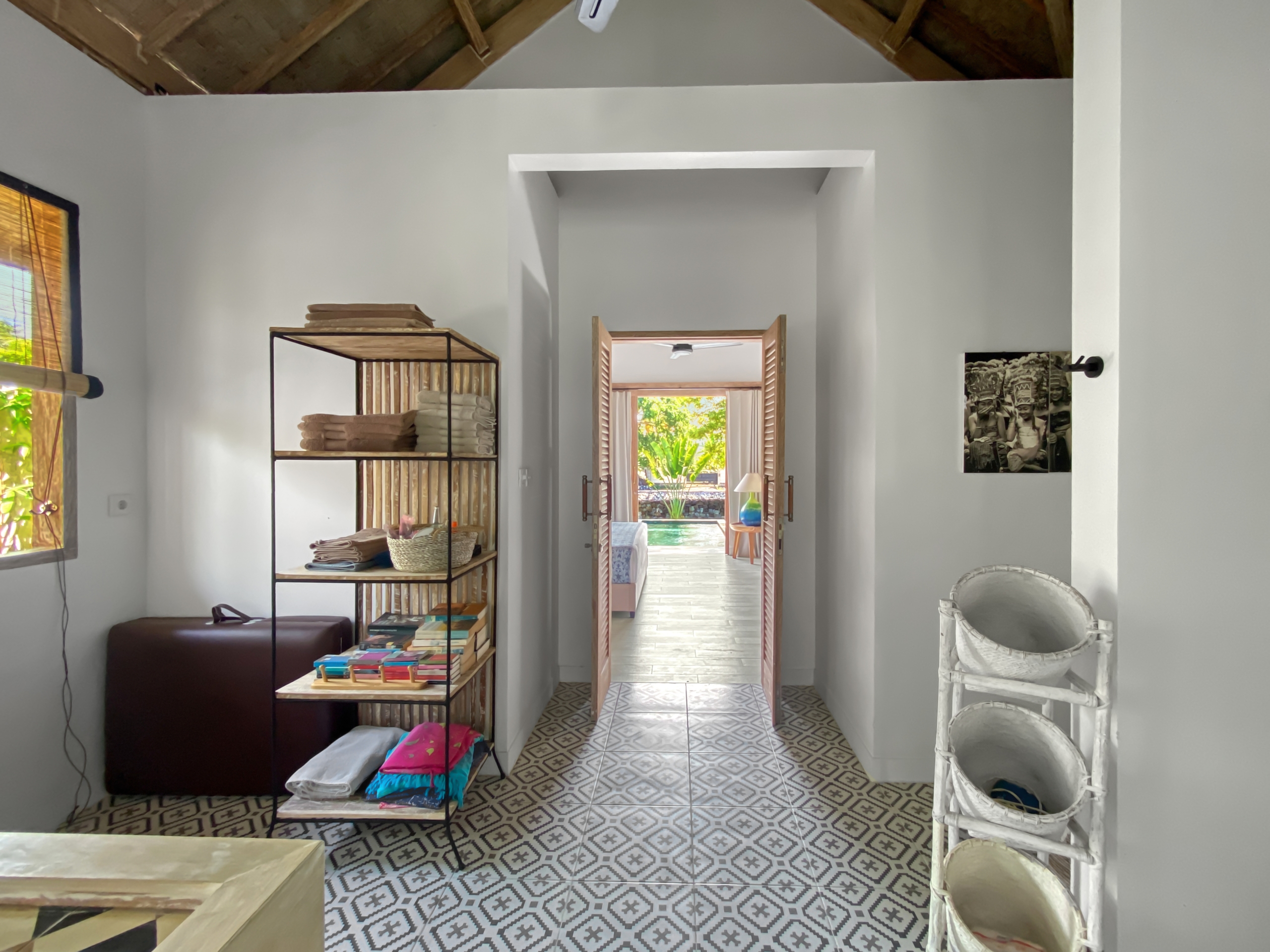 b8d8e732ae763aecf84f87bd50aabf1837213c67 Interior view of a bright room with a metal shelving unit holding towels and fabrics, leading to a sunlit doorway to an outdoor pool area.