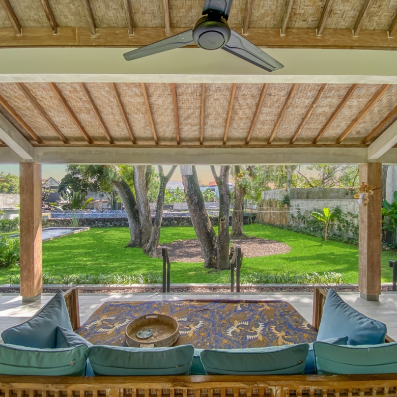 Covered outdoor seating area with a woven palm ceiling, wooden beams, and a ceiling fan overlooking a green garden with trees and plants.