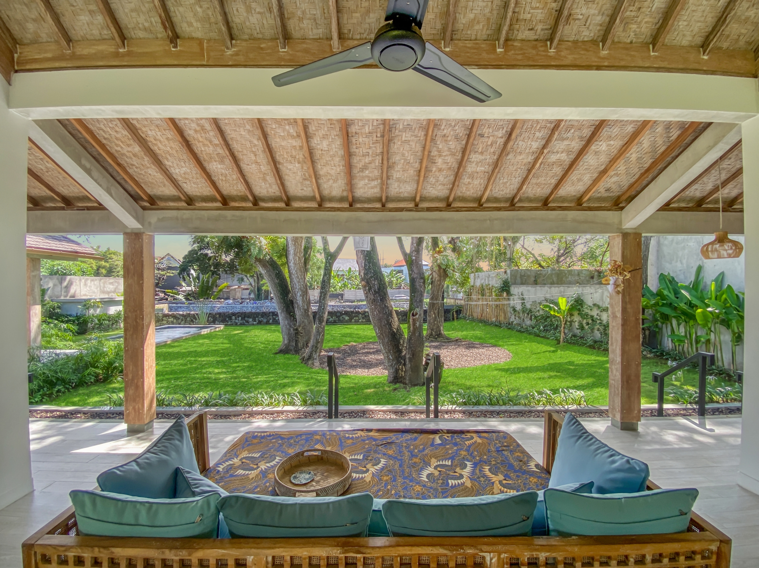 b9304f65926b3d7599584070460a2168f8de4035 Covered outdoor seating area with a woven palm ceiling, wooden beams, and a ceiling fan overlooking a green garden with trees and plants.