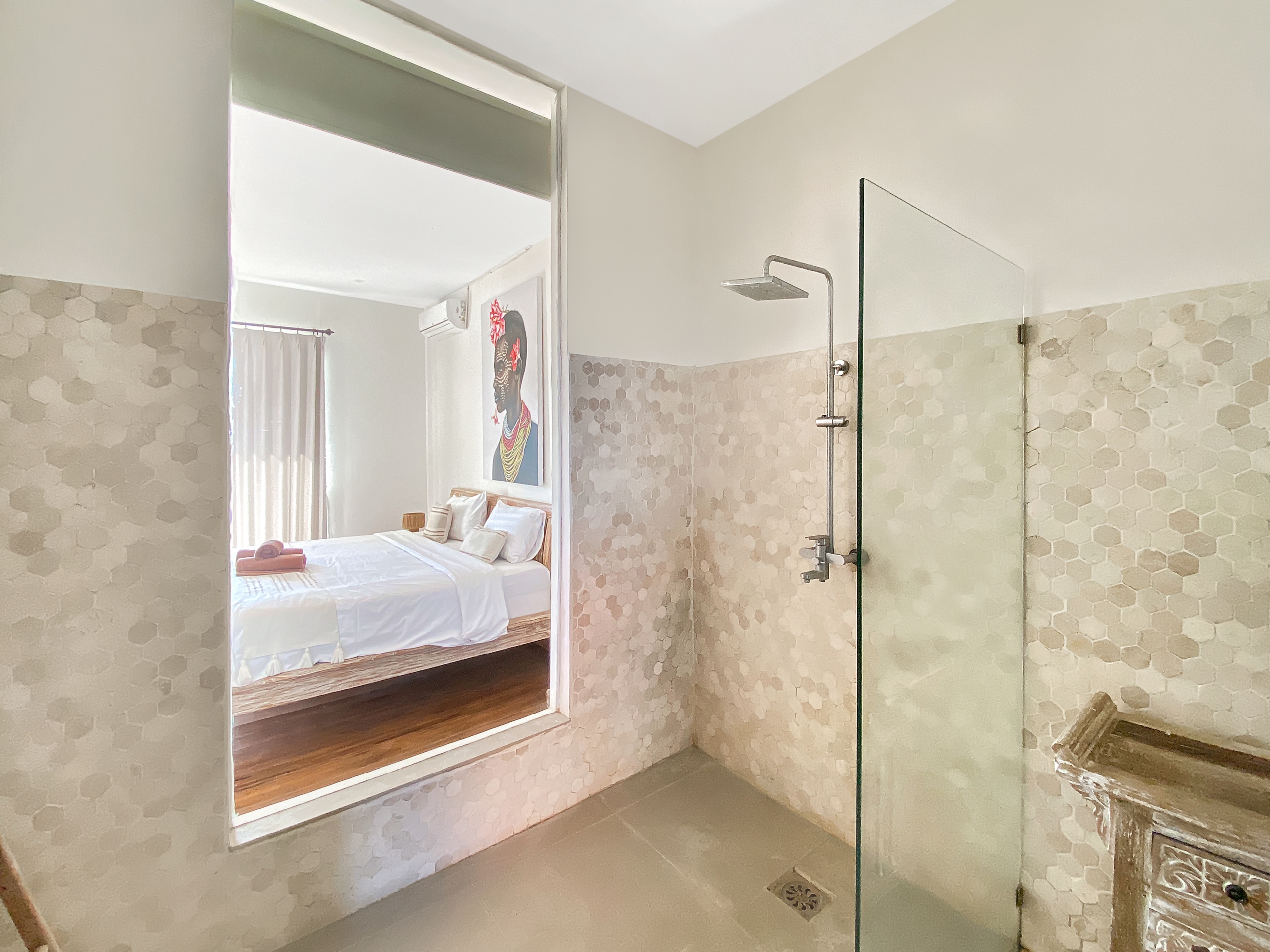 Bathroom with glass-enclosed shower and hexagonal tile walls leading to a bedroom through an open doorway. A rain shower head is visible on the right.