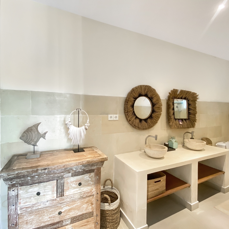 Showroom bathroom display: beige tiled wall with three round wicker-framed mirrors above white sinks holding stone bowls; a weathered wooden cabinet on the left and wicker baskets nearby.