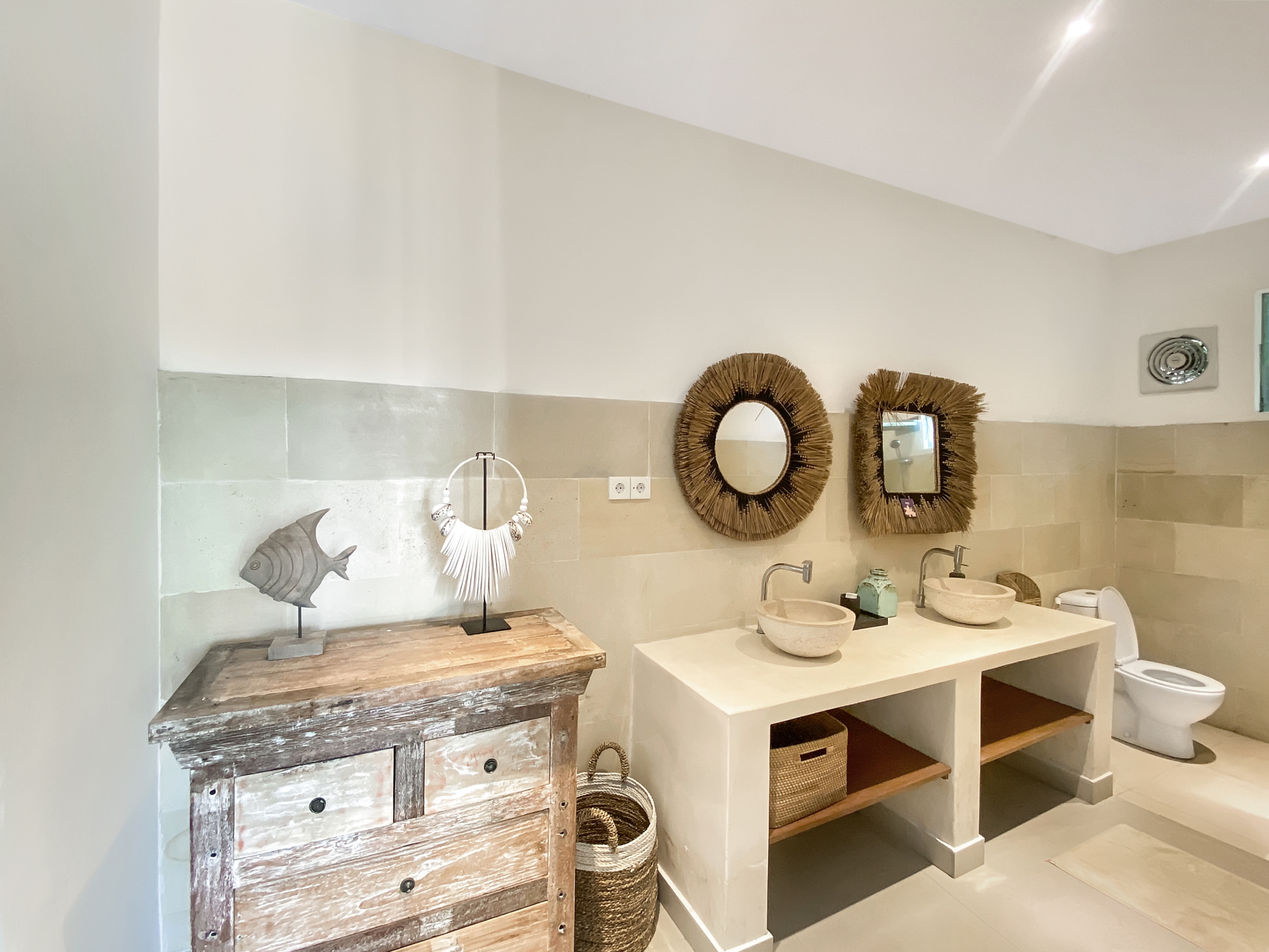 Showroom bathroom display: beige tiled wall with three round wicker-framed mirrors above white sinks holding stone bowls; a weathered wooden cabinet on the left and wicker baskets nearby.