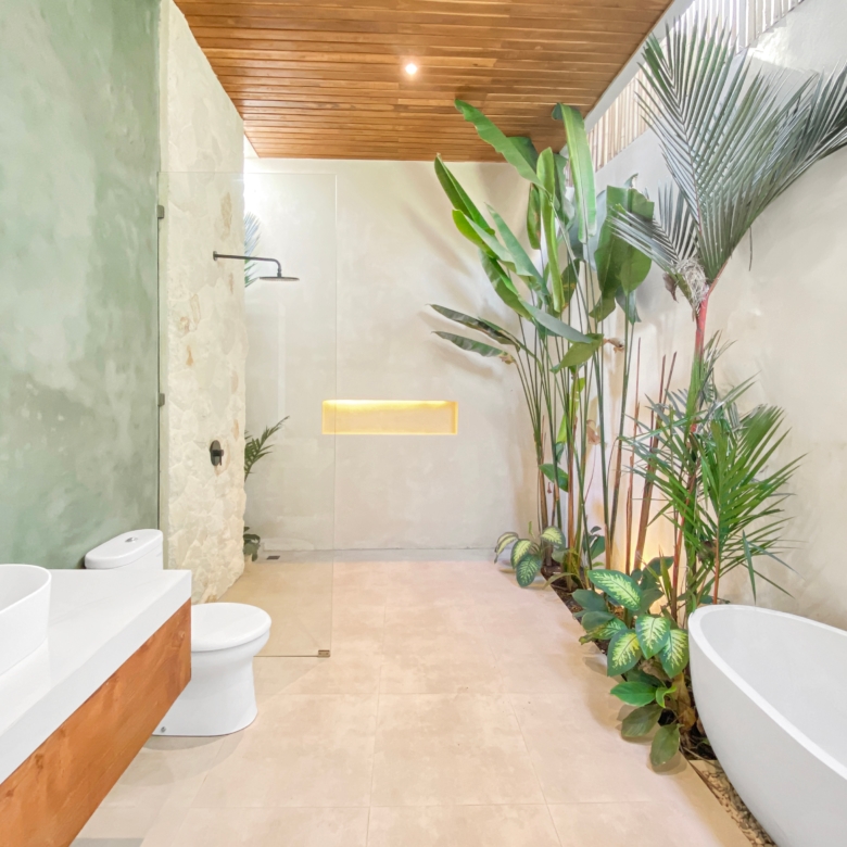 Spa-like bathroom with a freestanding white tub, abundant tropical plants along the right, beige tiled floor, and a wooden ceiling. On the left, a glass-walled shower area with a stone wall.