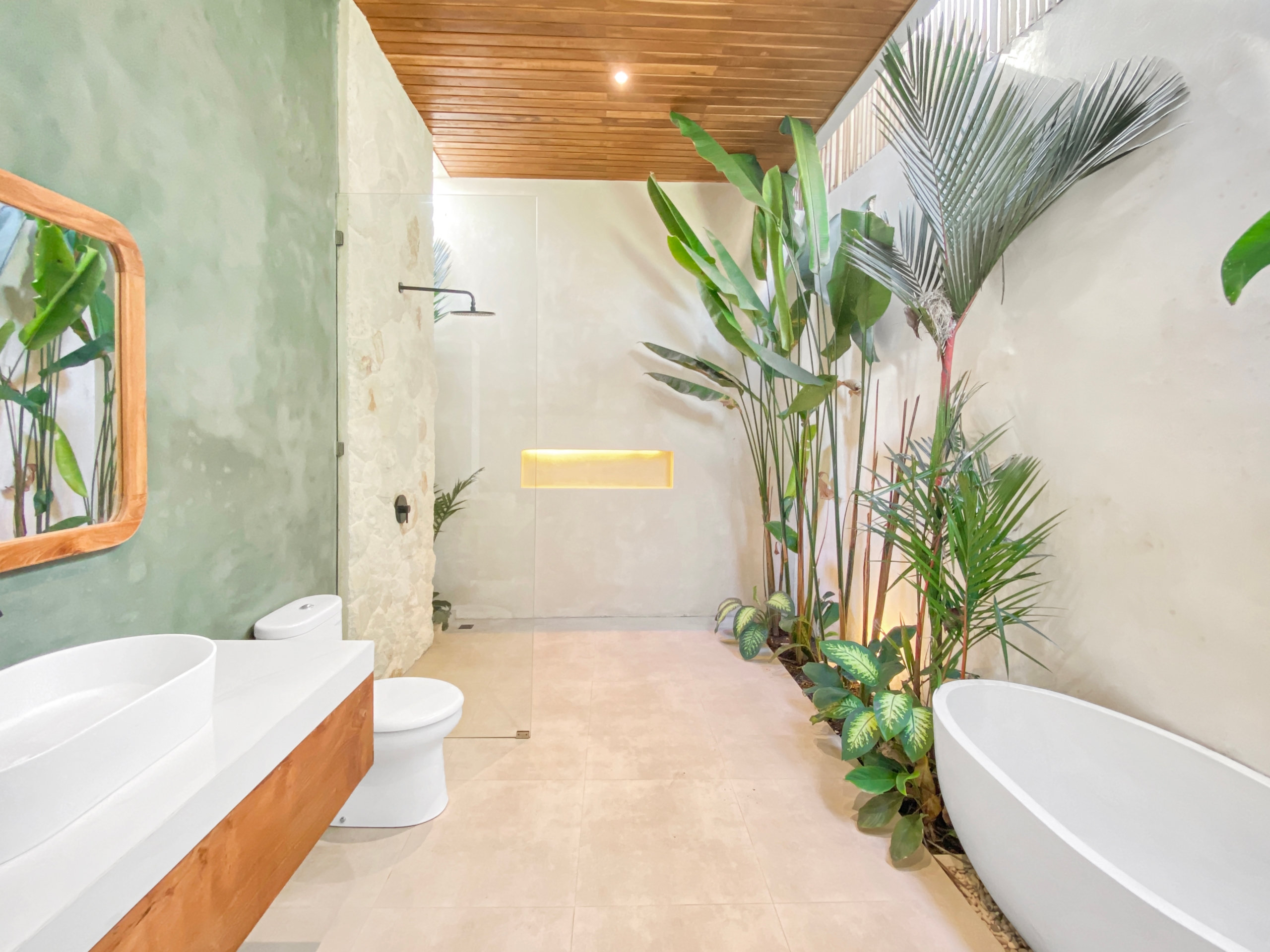 Spa-like bathroom with a freestanding white tub, abundant tropical plants along the right, beige tiled floor, and a wooden ceiling. On the left, a glass-walled shower area with a stone wall.