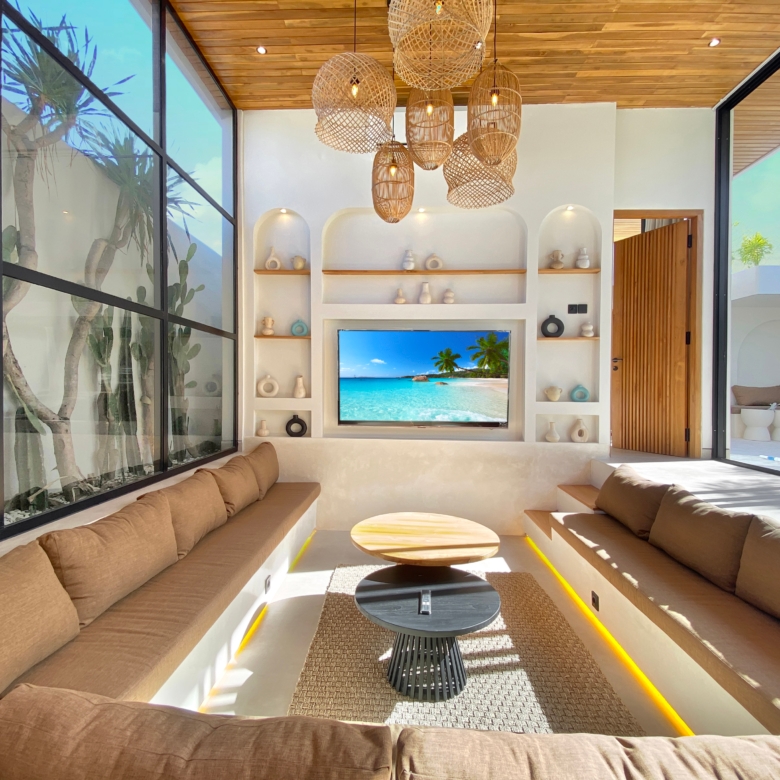 Sunlit living room with beige sectional sofas, round wooden coffee tables, and a tropical beach on the TV screen.