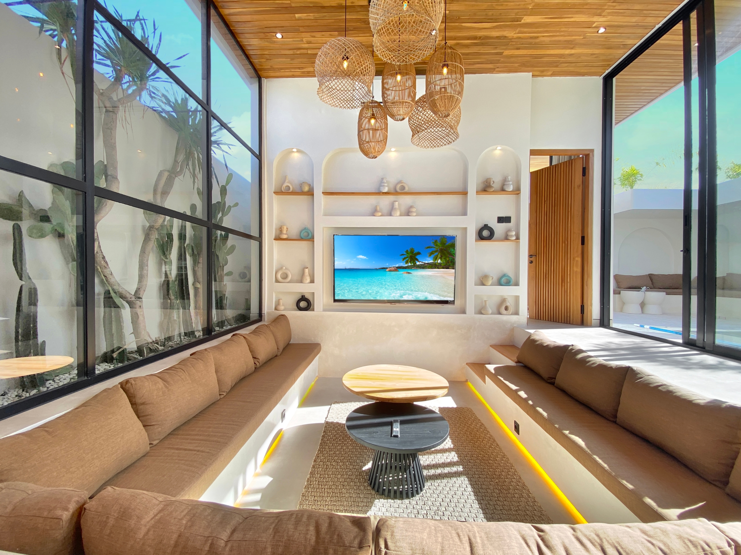 Sunlit living room with beige sectional sofas, round wooden coffee tables, and a tropical beach on the TV screen.