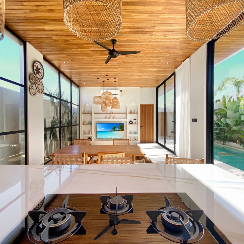 Open-concept kitchen and dining area with a gas stove island in the foreground, glass walls, and a wooden ceiling.