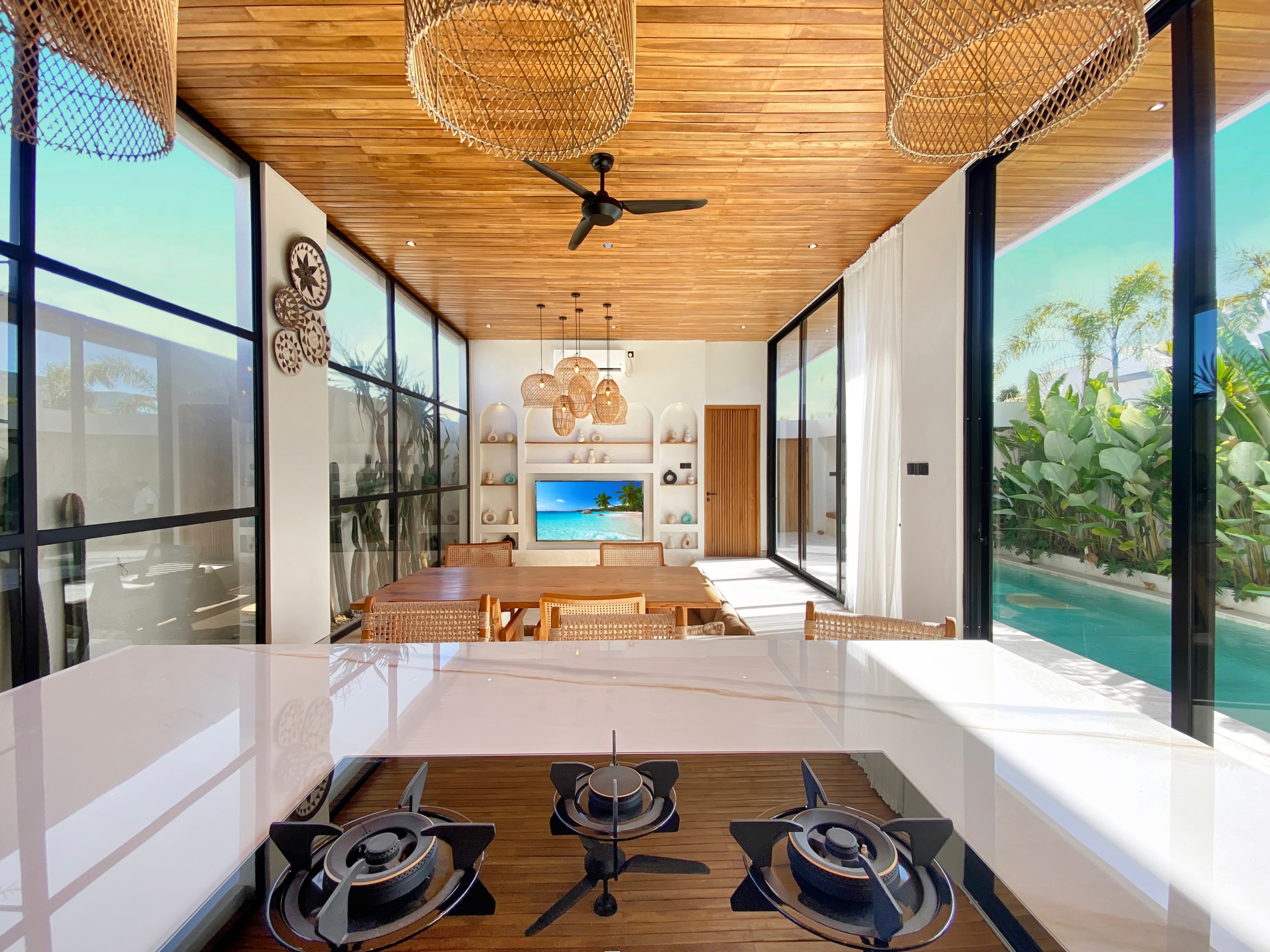 Open-concept kitchen and dining area with a gas stove island in the foreground, glass walls, and a wooden ceiling.