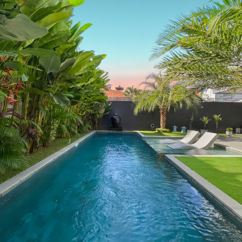 Rectangular pool bordered by tropical plants and a seating area with white lounge chairs in a sunny backyard.