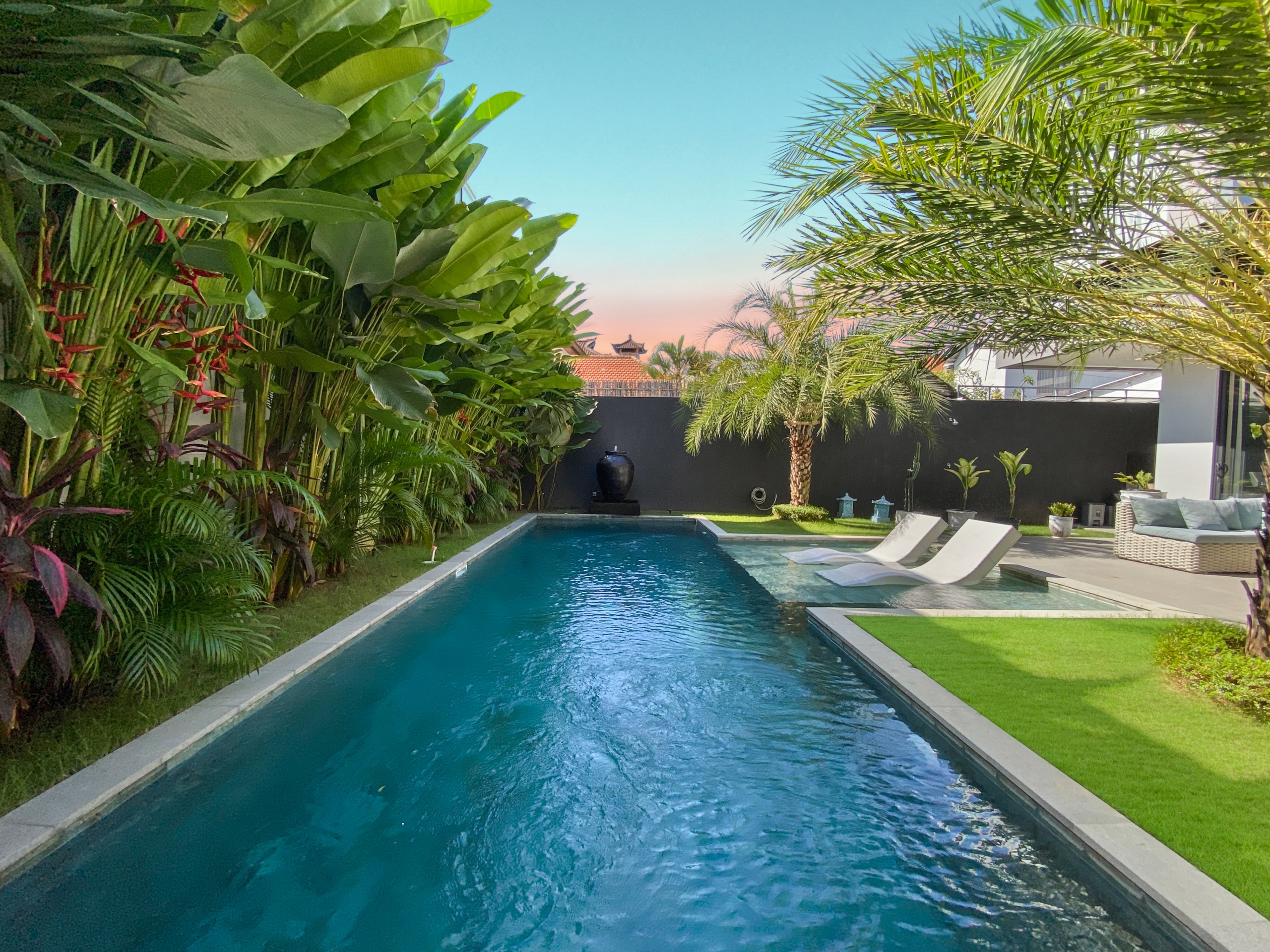 Rectangular pool bordered by tropical plants and a seating area with white lounge chairs in a sunny backyard.