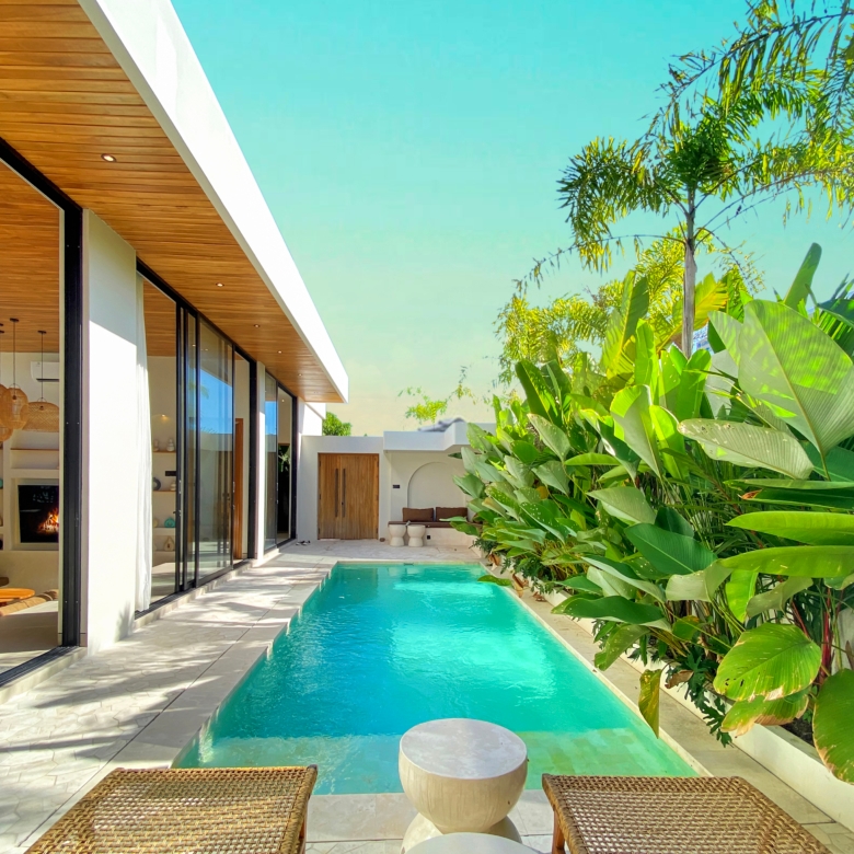 Modern house with glass sliding doors, wooden overhang, and a turquoise pool bordered by tropical plants.