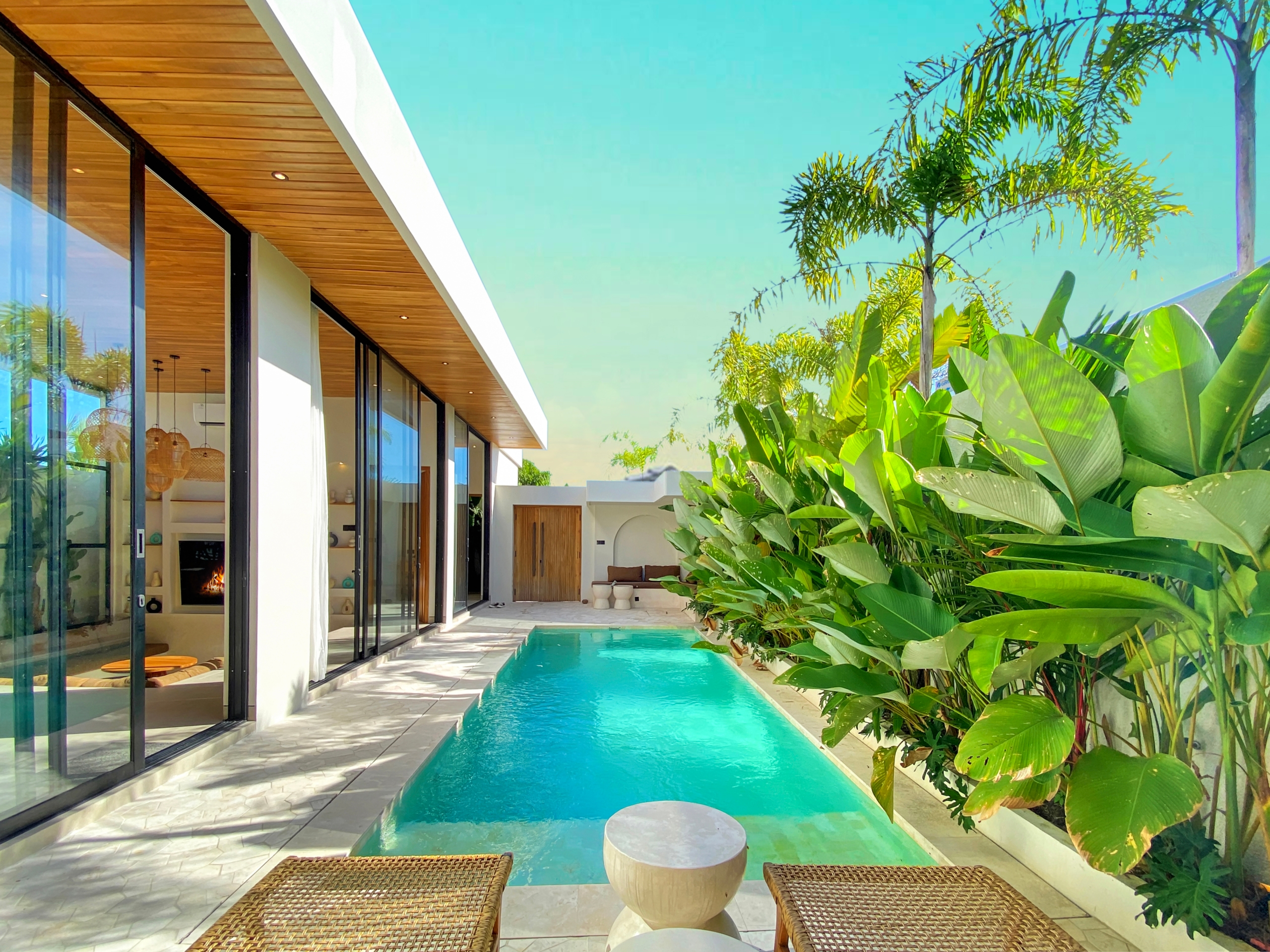 Modern house with glass sliding doors, wooden overhang, and a turquoise pool bordered by tropical plants.