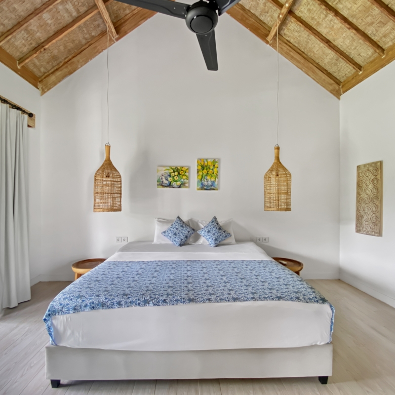 Bright bedroom with a large bed in white linens and a blue patterned throw, plus wicker pendant lamps and a thatched ceiling fan.