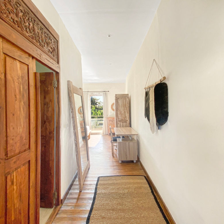 Long narrow hallway with a carved wooden door frame on the left and a sunlit view at the far end.