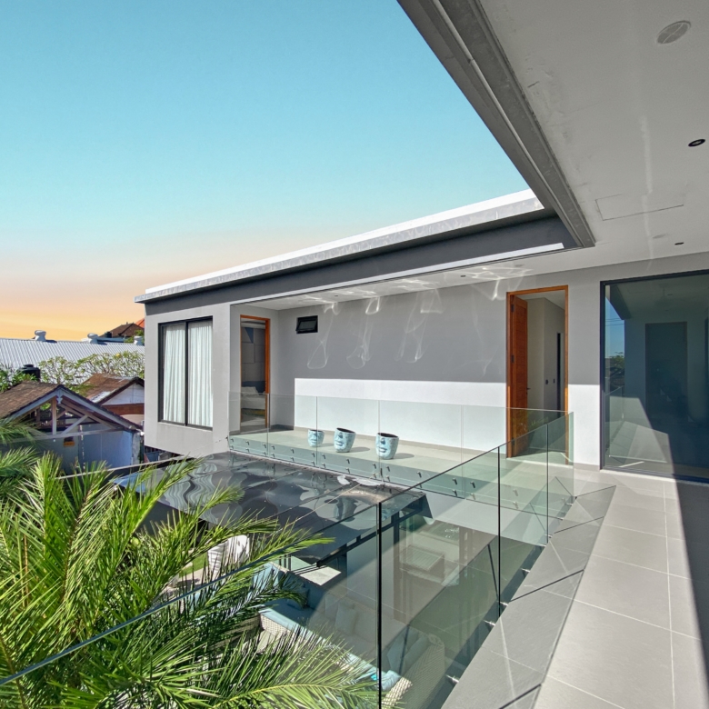 Modern house exterior with a glass-railed balcony, gray tiled patio, tropical plants, and open doors under a clear blue sky.