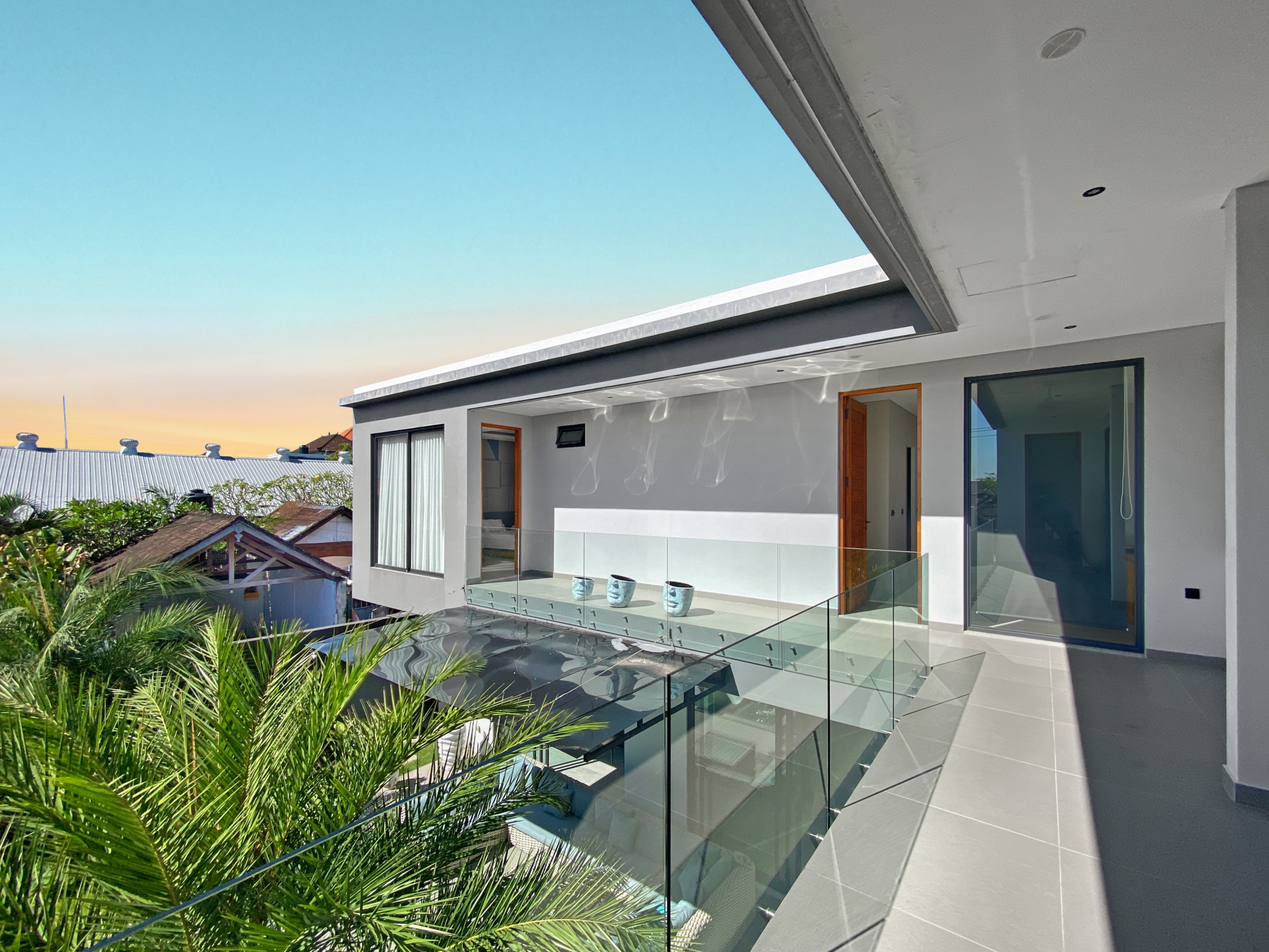 Modern house exterior with a glass-railed balcony, gray tiled patio, tropical plants, and open doors under a clear blue sky.