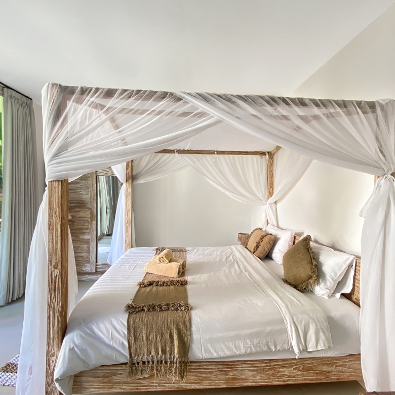 Four-poster bed with white sheer canopy and brown accents in a bright tropical-style bedroom.