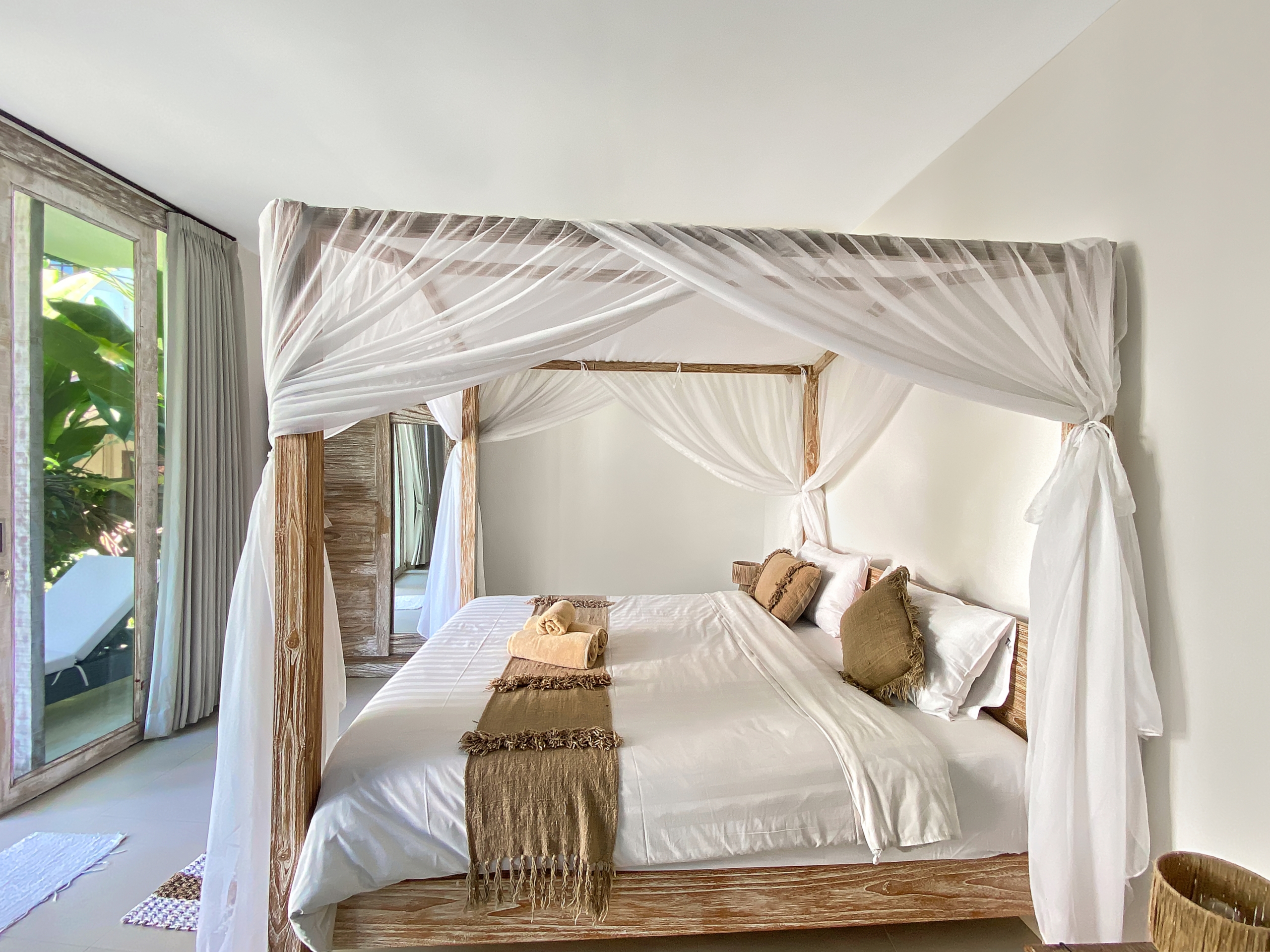 Four-poster bed with white sheer canopy and brown accents in a bright tropical-style bedroom.