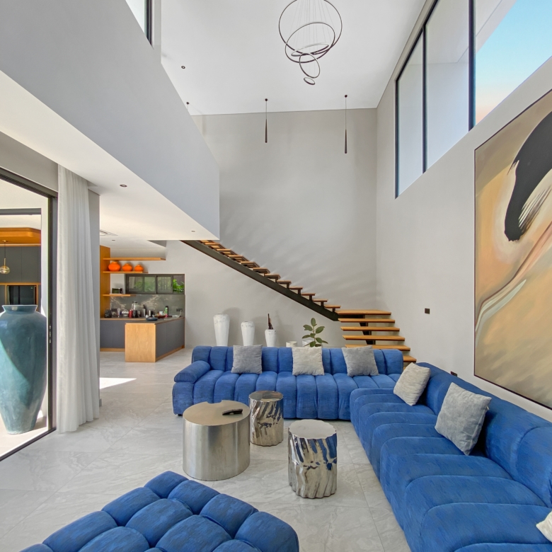 Open-concept living room with a bright, spacious feel: blue sectional sofa, round metal coffee tables, and a tall staircase leading to a second level, plus a large mural on the right wall.