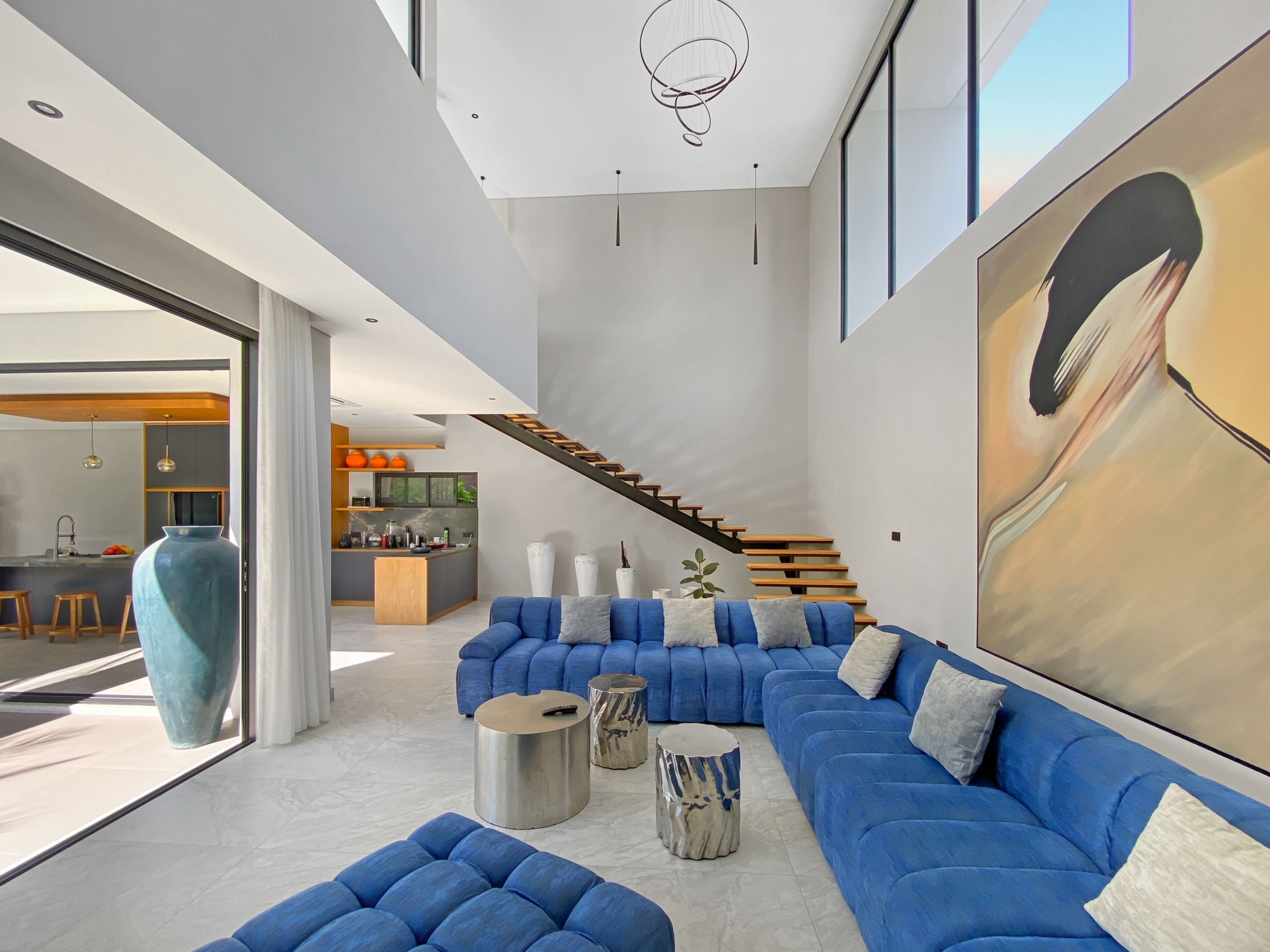Open-concept living room with a bright, spacious feel: blue sectional sofa, round metal coffee tables, and a tall staircase leading to a second level, plus a large mural on the right wall.