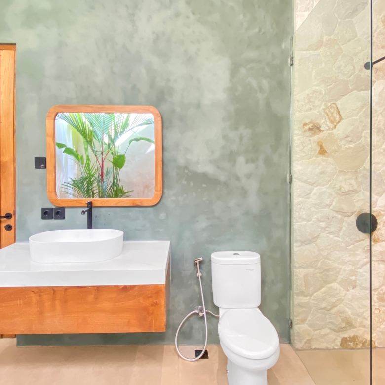 Modern bathroom with wooden cabinet, green plaster wall, round sink, mirror, and glass shower with stone wall.