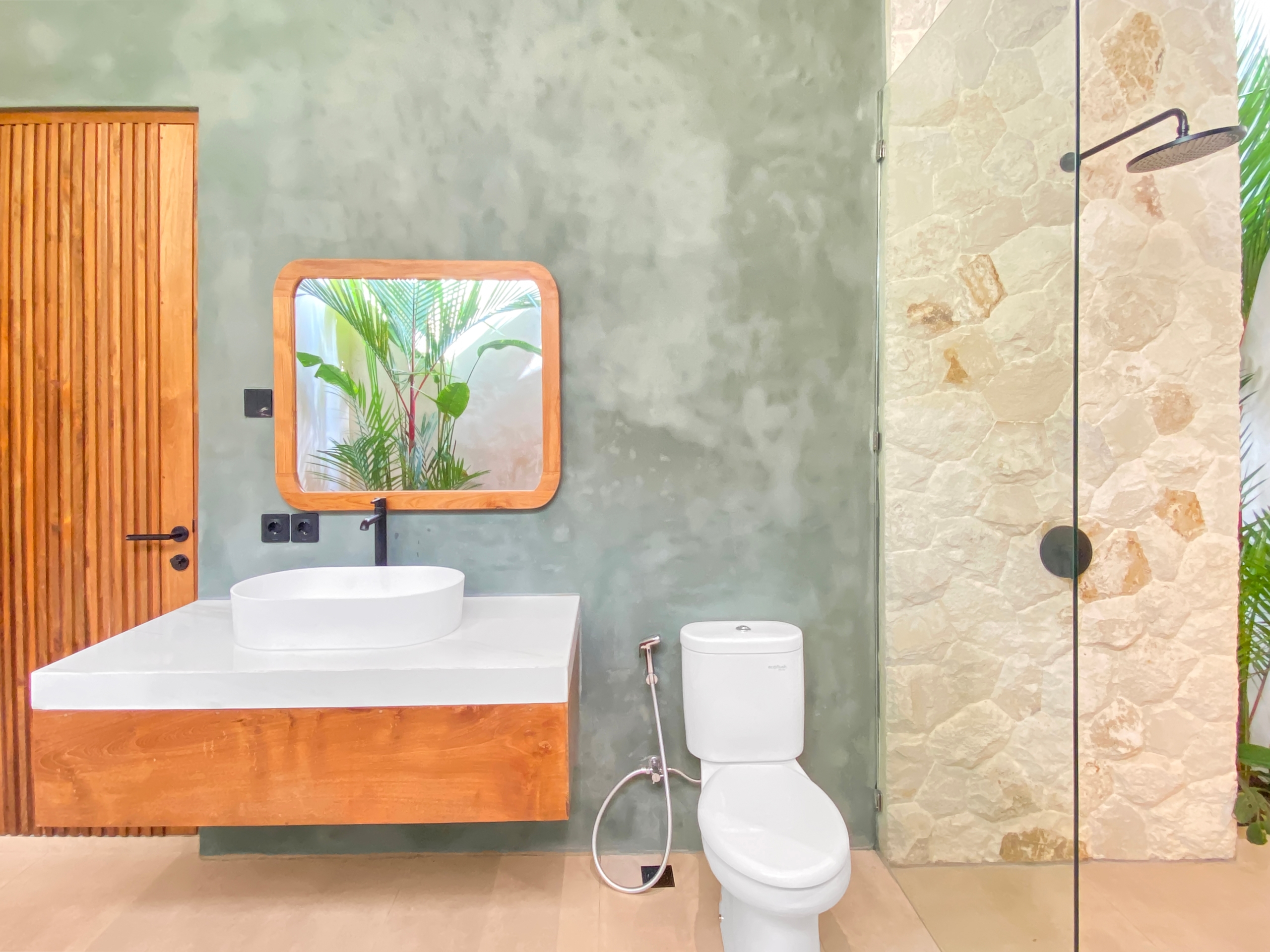 Modern bathroom with wooden cabinet, green plaster wall, round sink, mirror, and glass shower with stone wall.