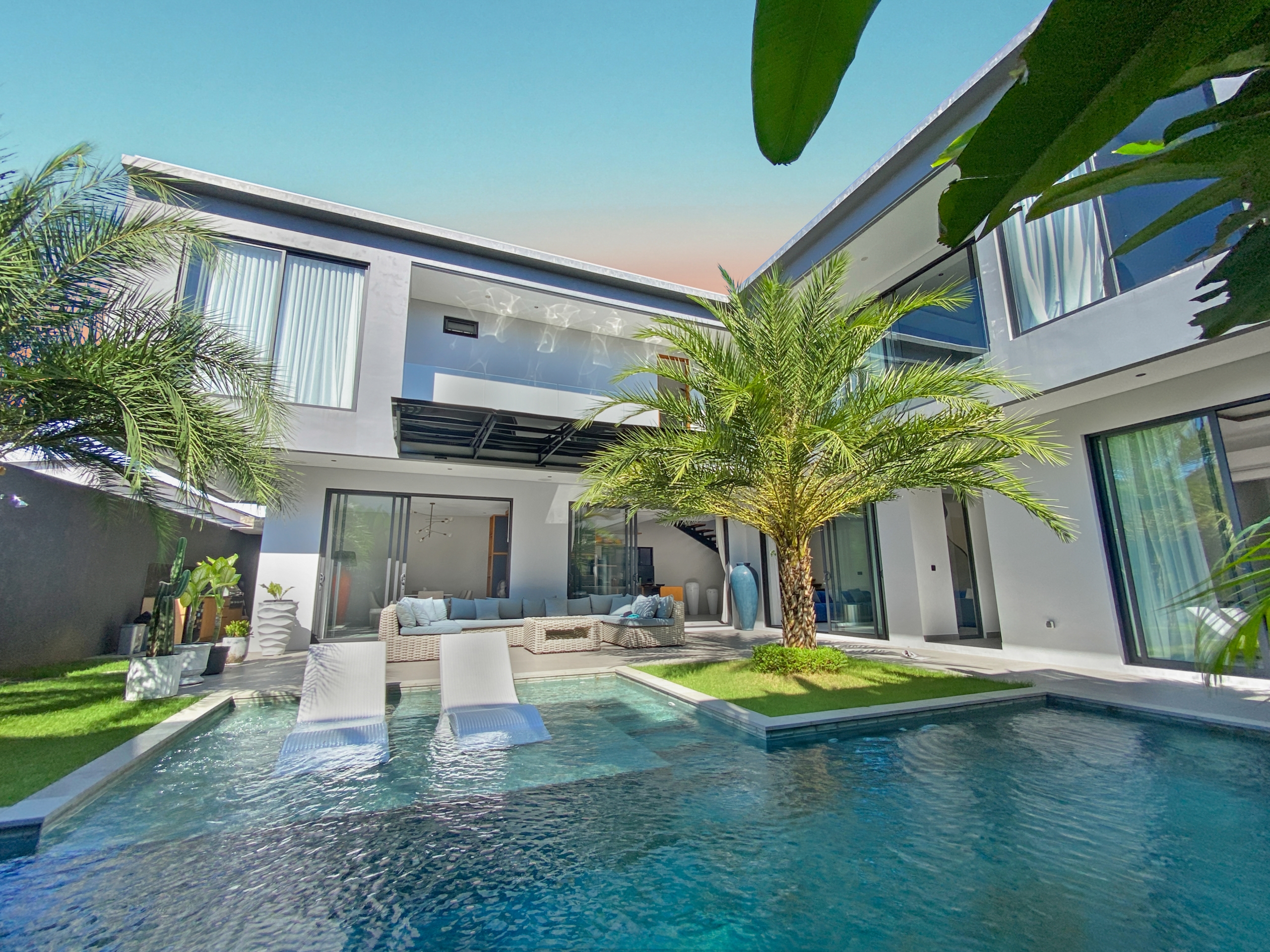 Modern two-story white house with glass doors, palm trees, and a rectangular pool with white loungers outside.