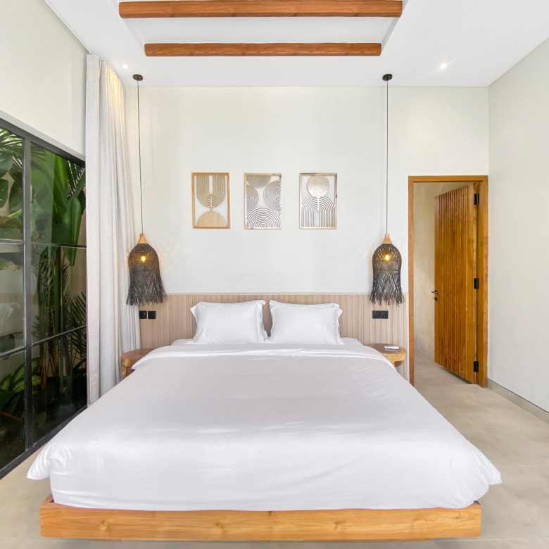 Modern hotel bedroom with a large white bed, wooden frame, and matching pendant lights; plants visible through a glass wall on the left.