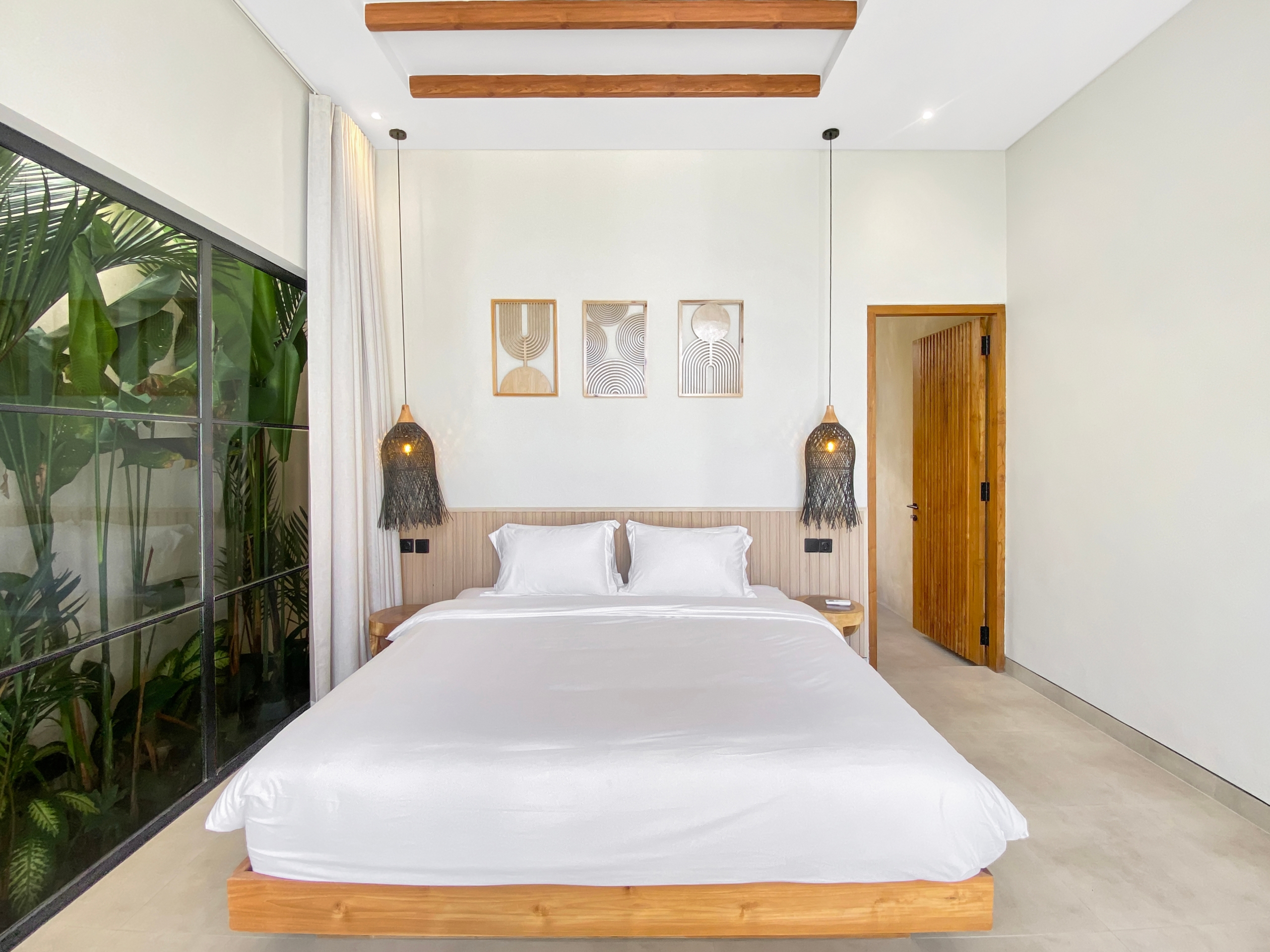 Modern hotel bedroom with a large white bed, wooden frame, and matching pendant lights; plants visible through a glass wall on the left.