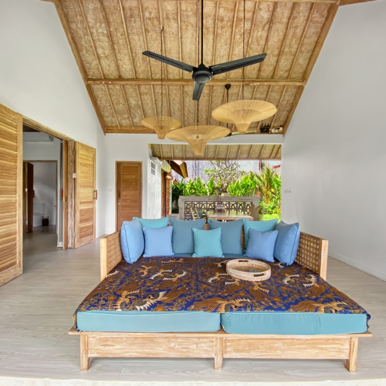 Open-air living area with a blue-cushioned wicker sofa under a wooden thatched ceiling and ceiling fans.