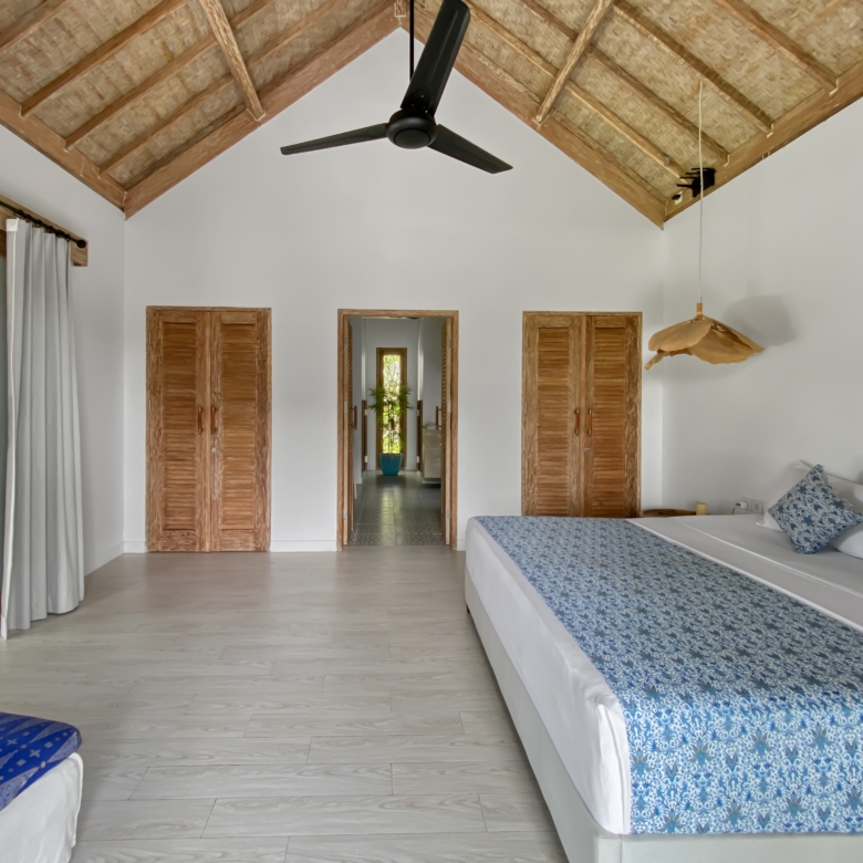 A bright hotel-style bedroom with a high thatched ceiling, central black ceiling fan, and two beds with blue patterned covers against white walls.