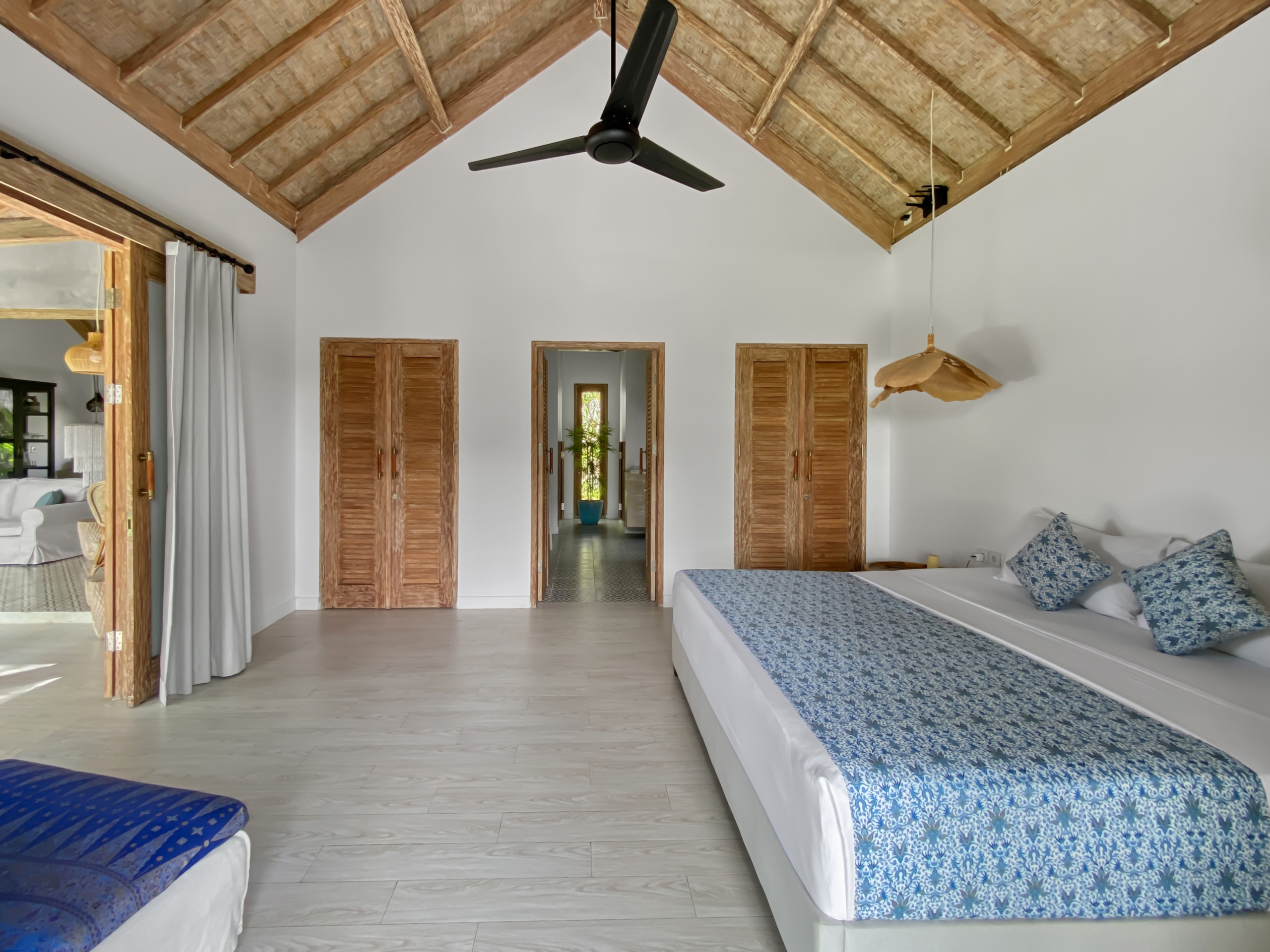 d80649d582bb9e0a010d54609d566921c2ee6884 A bright hotel-style bedroom with a high thatched ceiling, central black ceiling fan, and two beds with blue patterned covers against white walls.