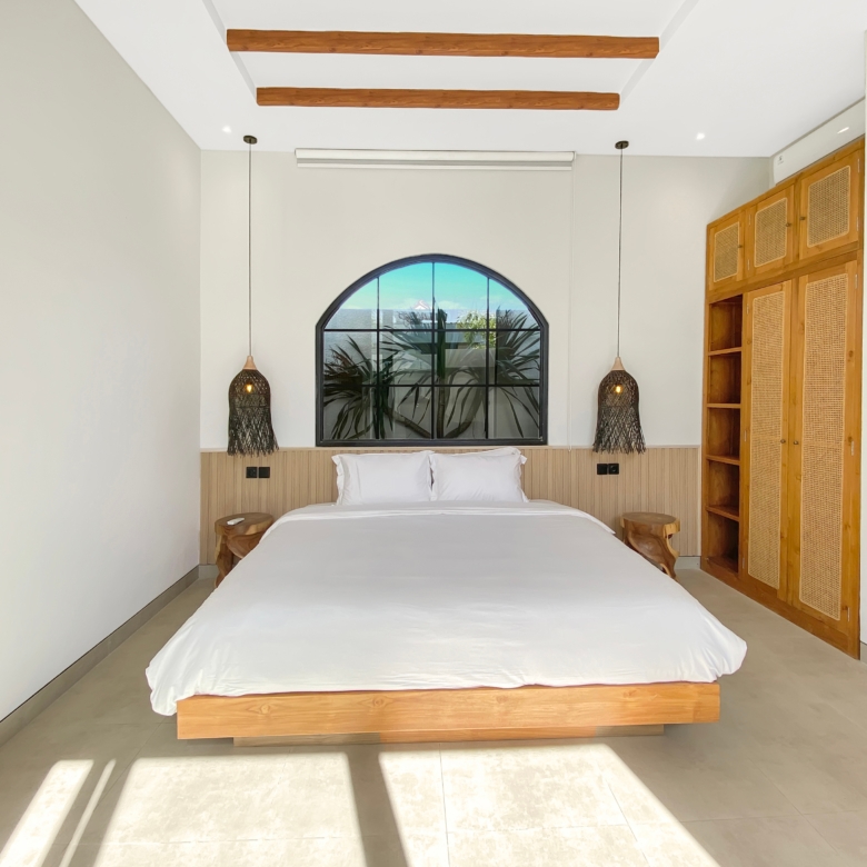 Hotel-style bedroom with low wooden bed, white bedding, and a large arched window behind the headboard. Two woven pendant lights hang on either side.