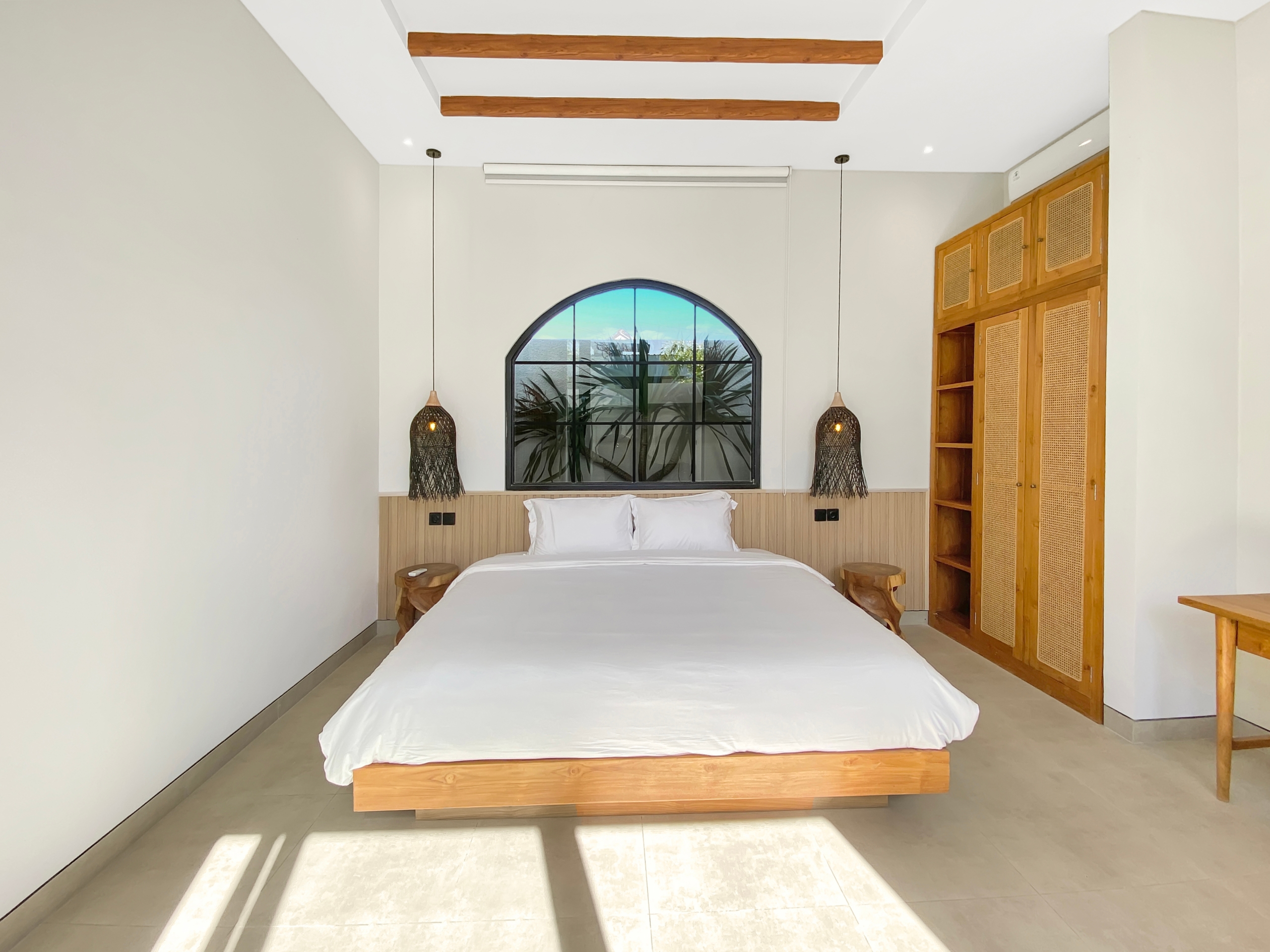 Hotel-style bedroom with low wooden bed, white bedding, and a large arched window behind the headboard. Two woven pendant lights hang on either side.