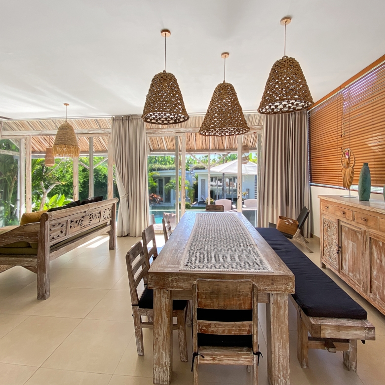 Bright tropical dining room featuring a long rustic wooden table, woven pendant lights, and large windows with blinds.