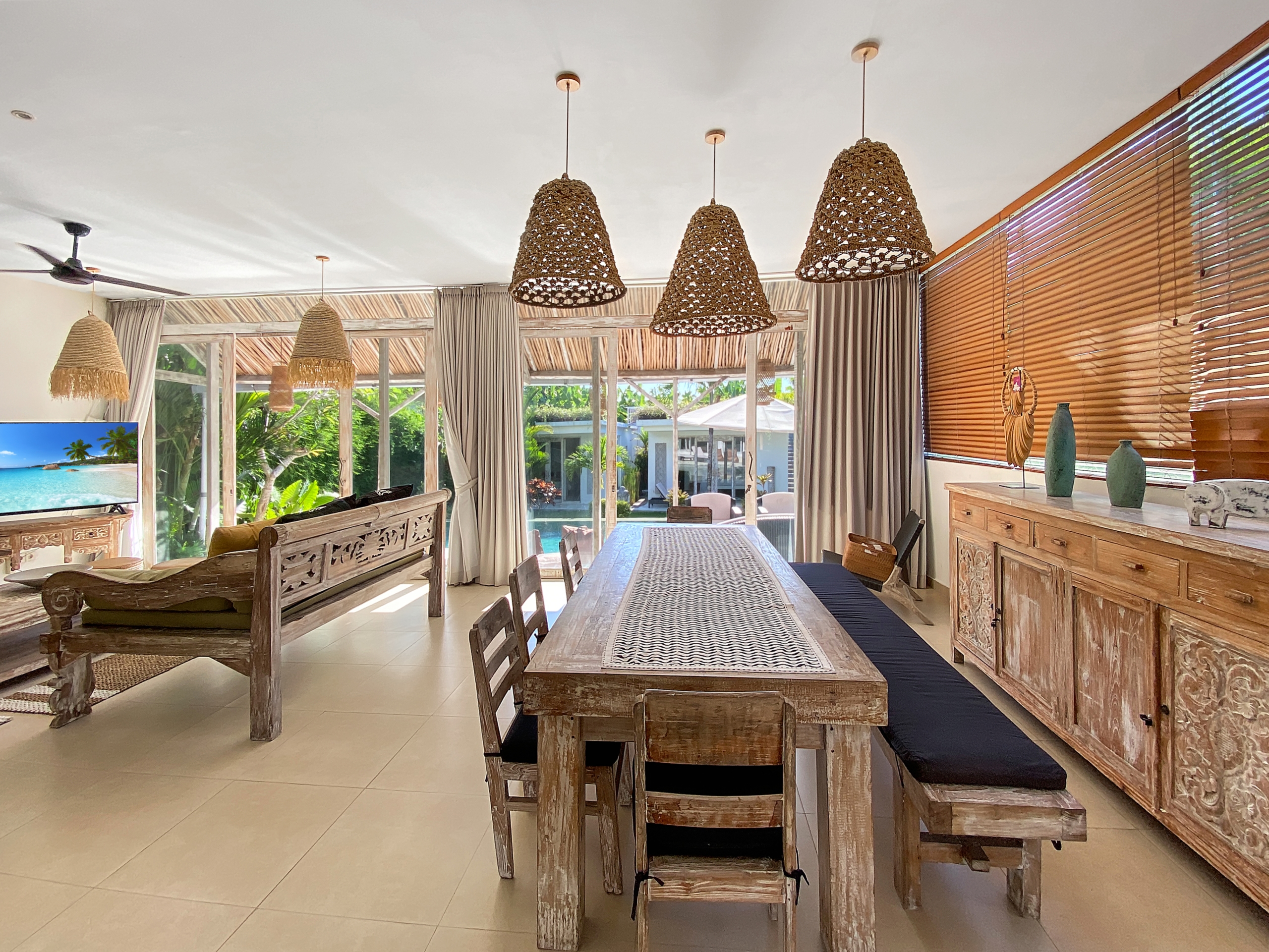 Bright tropical dining room featuring a long rustic wooden table, woven pendant lights, and large windows with blinds.