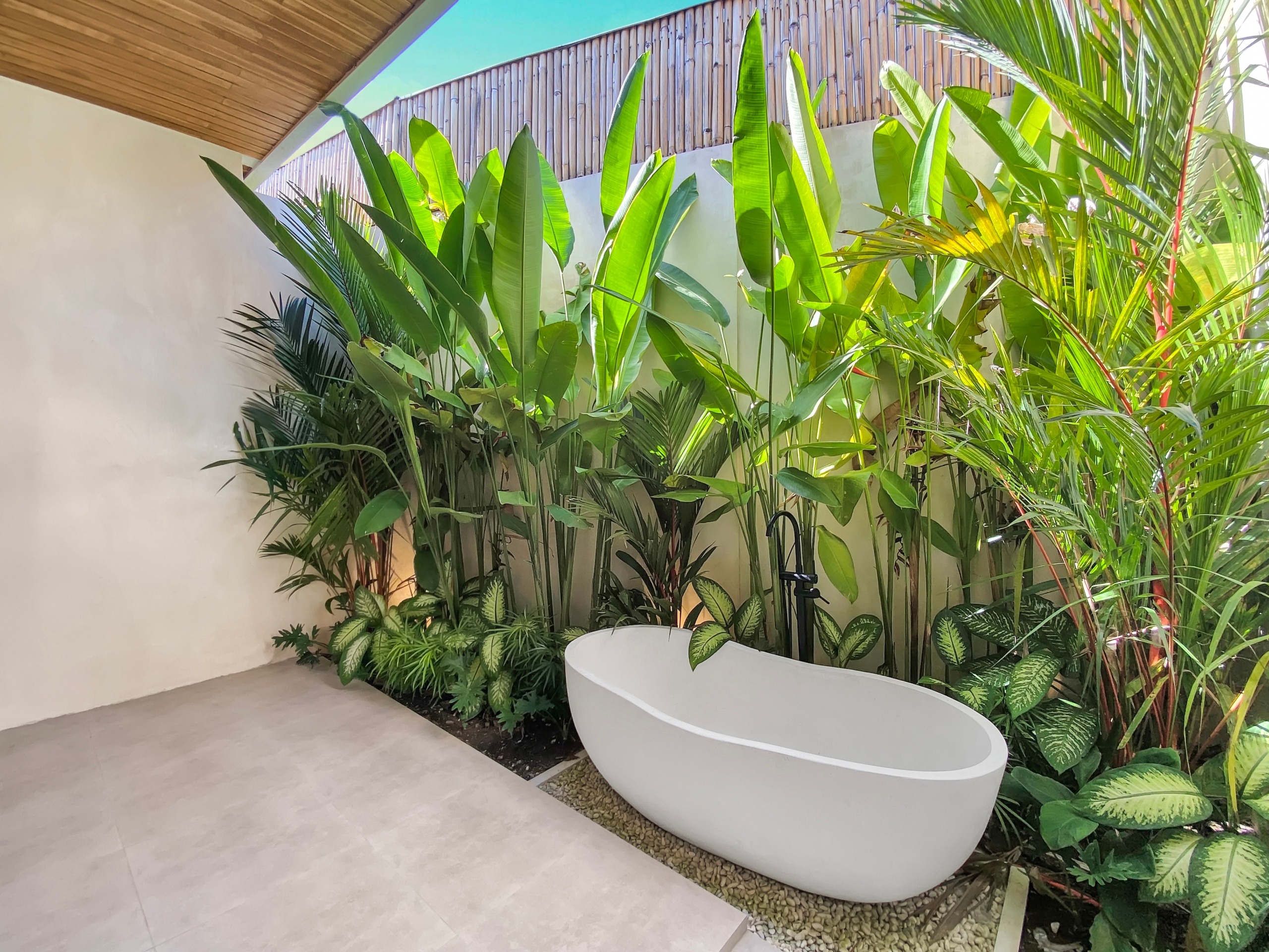 Indoor courtyard with a curved white planter filled with tall tropical plants along a beige wall and light tile floor, under a wooden ceiling.