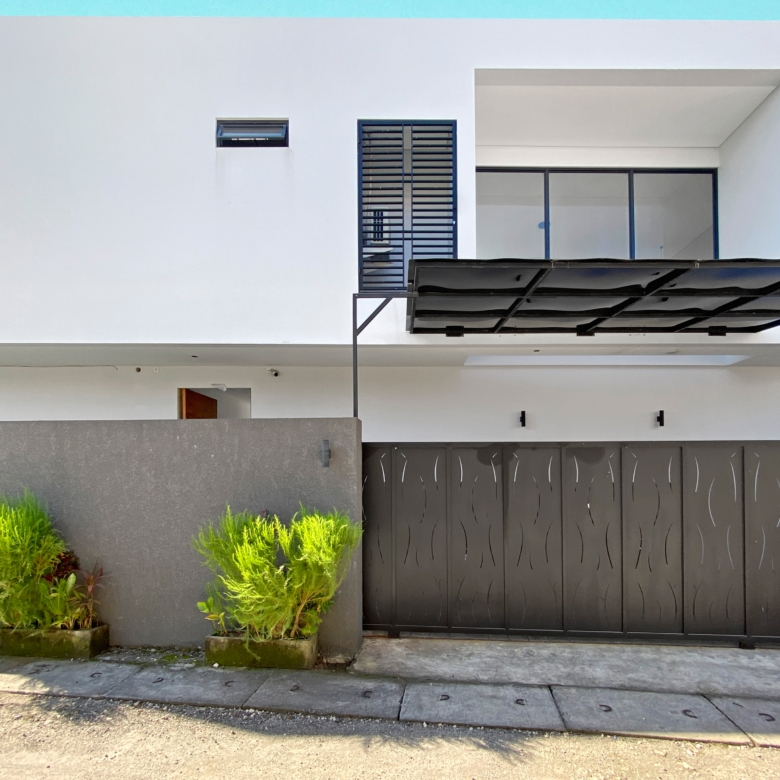 Modern white townhouse facade with a blue trim along the top, a balcony with glass doors, and a large black gate, plus green shrubs in front.