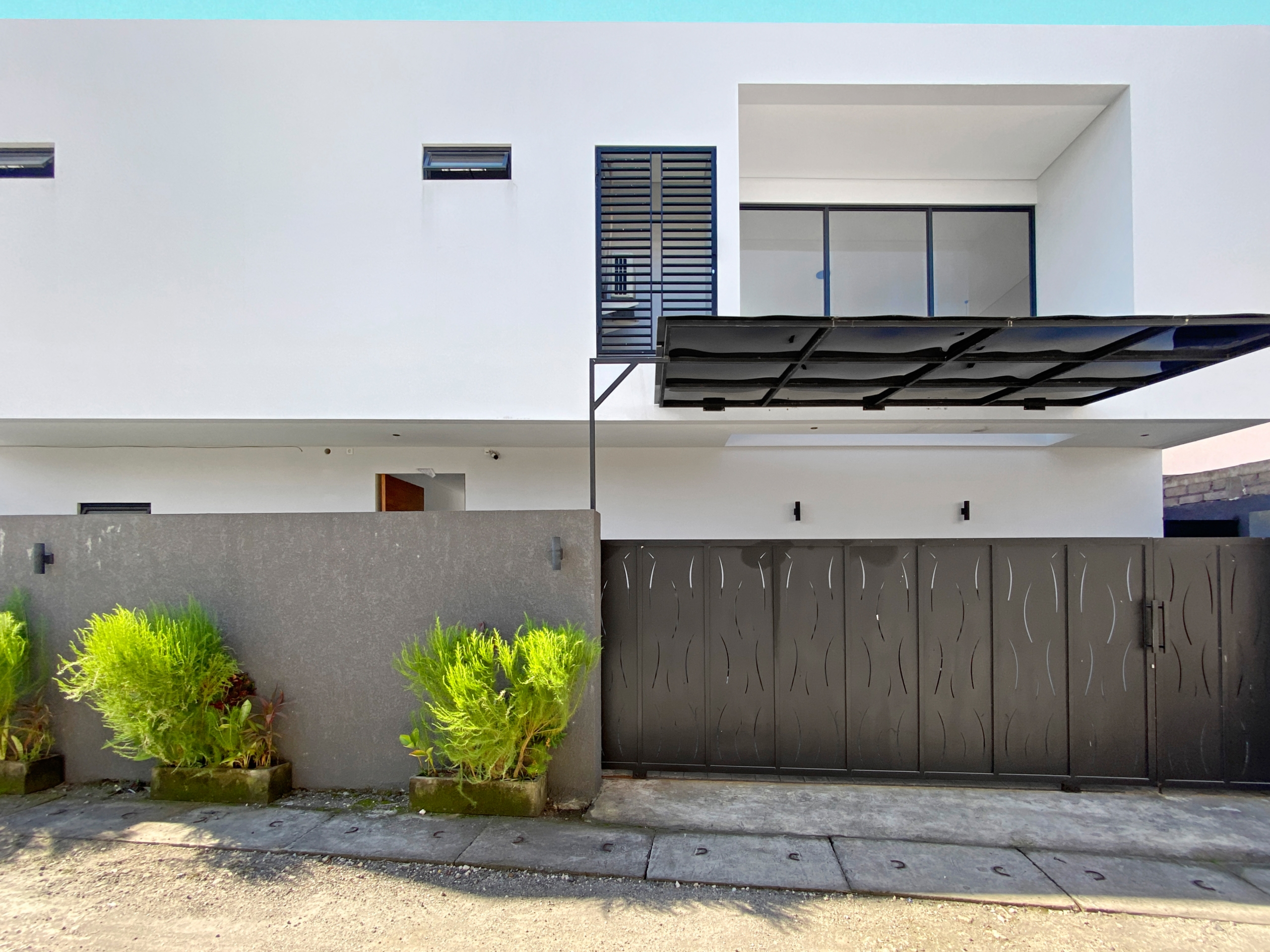 Modern white townhouse facade with a blue trim along the top, a balcony with glass doors, and a large black gate, plus green shrubs in front.