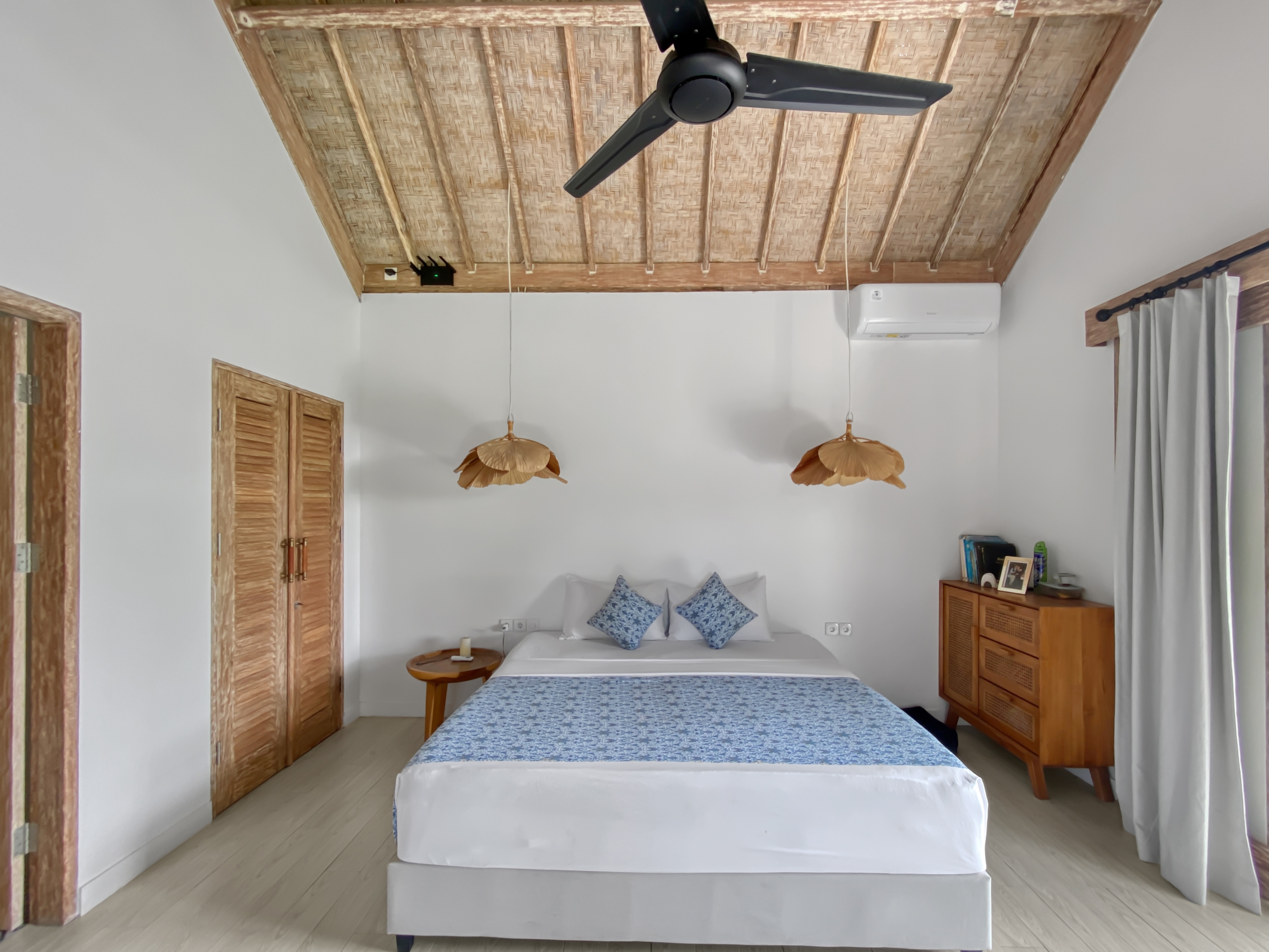 ec1cd04c5441d5acefbbe71db42c9cc632921c71 Bedroom with two twin beds pushed together, white bedding and blue patterned runners, and a wooden woven ceiling with a black ceiling fan.